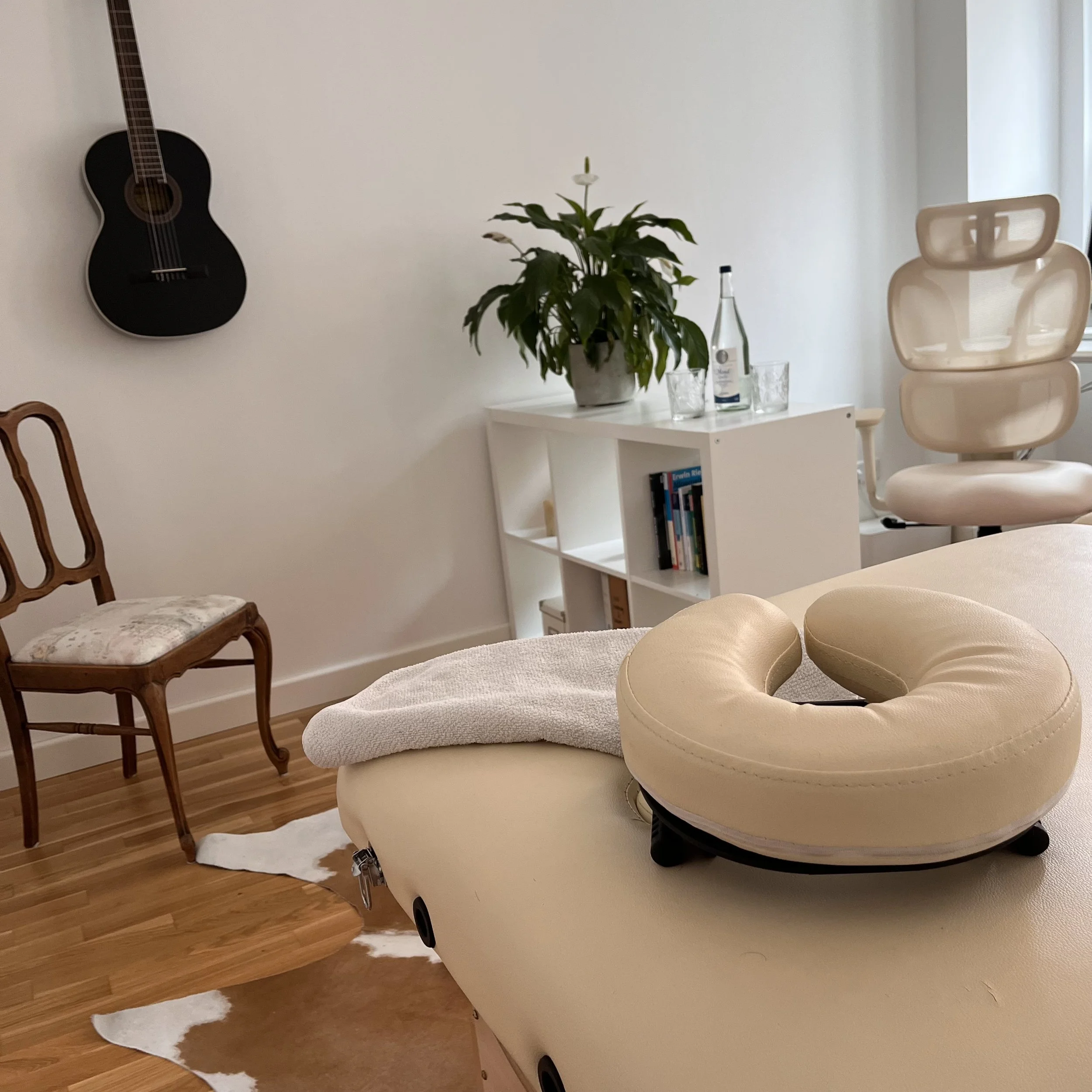 Gesundheits- oder Massagezimmer mit Behandlungsliege, Kopfstütze, holzockerngeschmiedetem Stuhl, weißem Bücherregal, Pflanzen, Wasserflasche, Gläser und einem bequemen weißen Anti-Dekubitus-Sitzkissen.