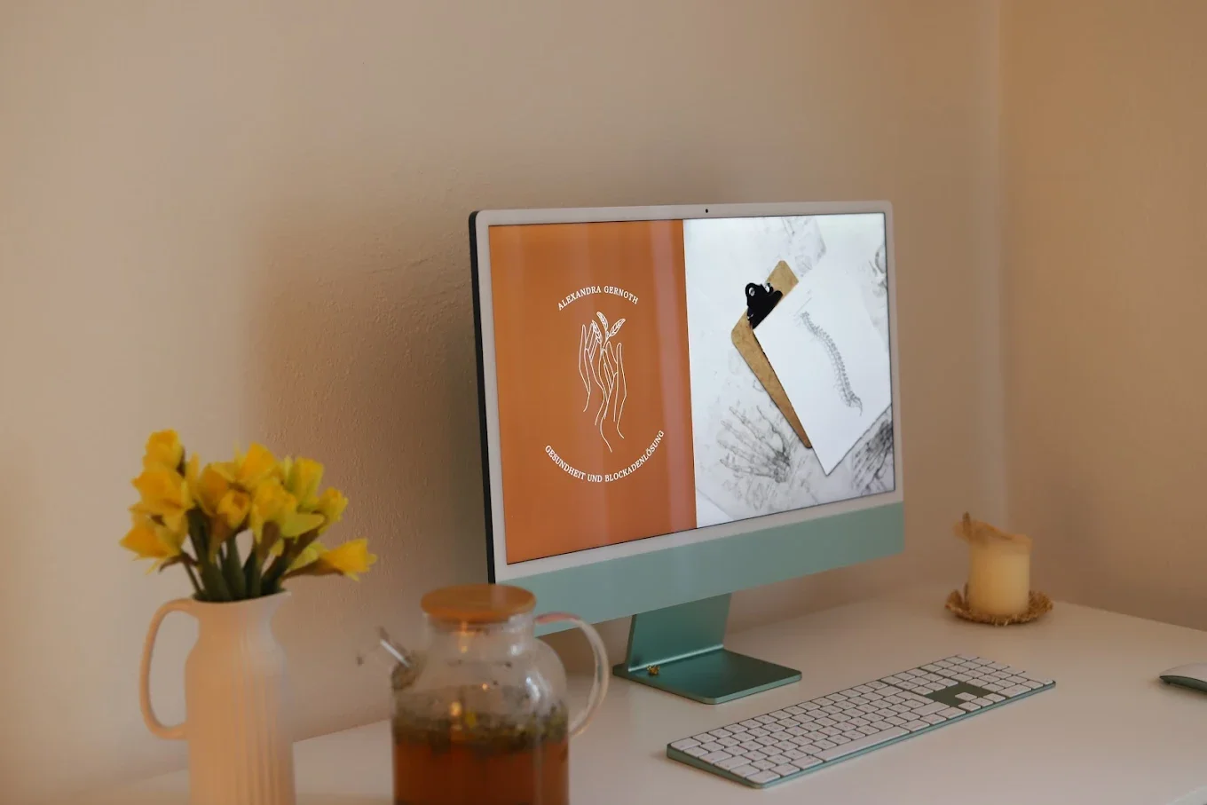 Ein Arbeitsplatz mit einem iMac-Computer, einer Tastatur, einer Vase mit gelben Blumen, einer Kerze und einem Glas mit Tee auf einem weißen Schreibtisch.