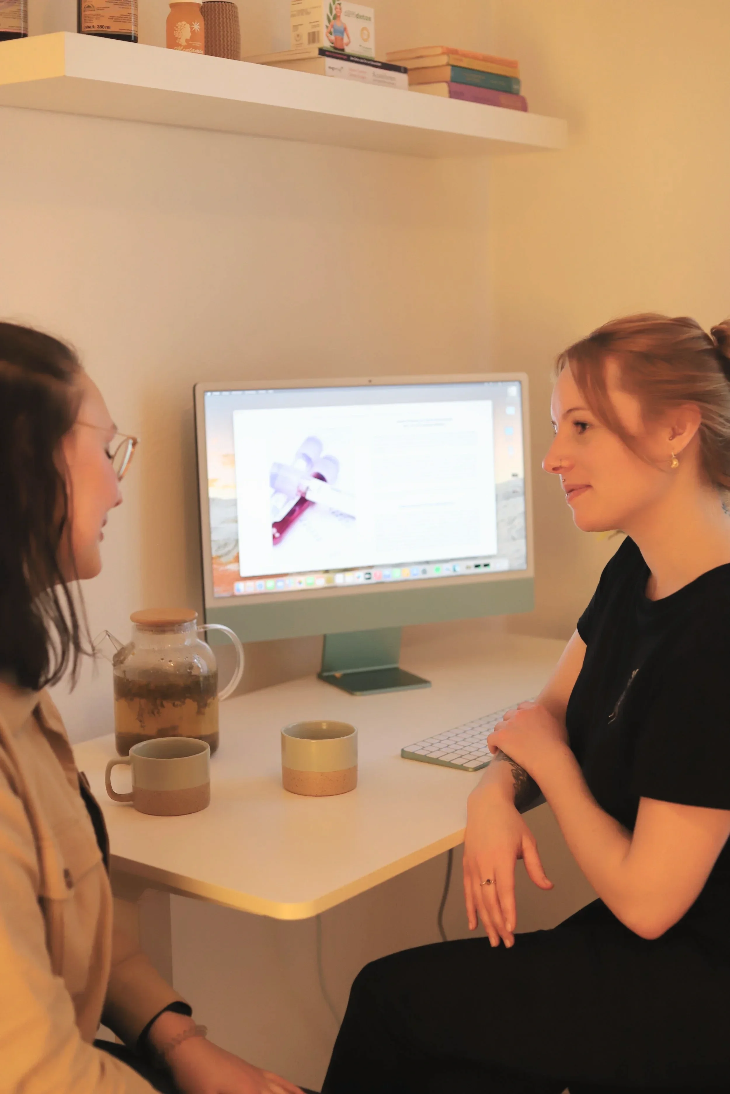 Zwei Frauen sitzen sich gegenüber an einem weißen Tisch in einem Raum mit weißen Wänden. Auf dem Tisch stehen zwei Tassen, eine Kanne mit Tee und eine Tastatur. Im Hintergrund ein Computerbildschirm, der eine Webseite oder einen Text anzeigt. Oben an der Wand befindet sich ein Regal mit bunten Büchern und anderen Gegenständen.