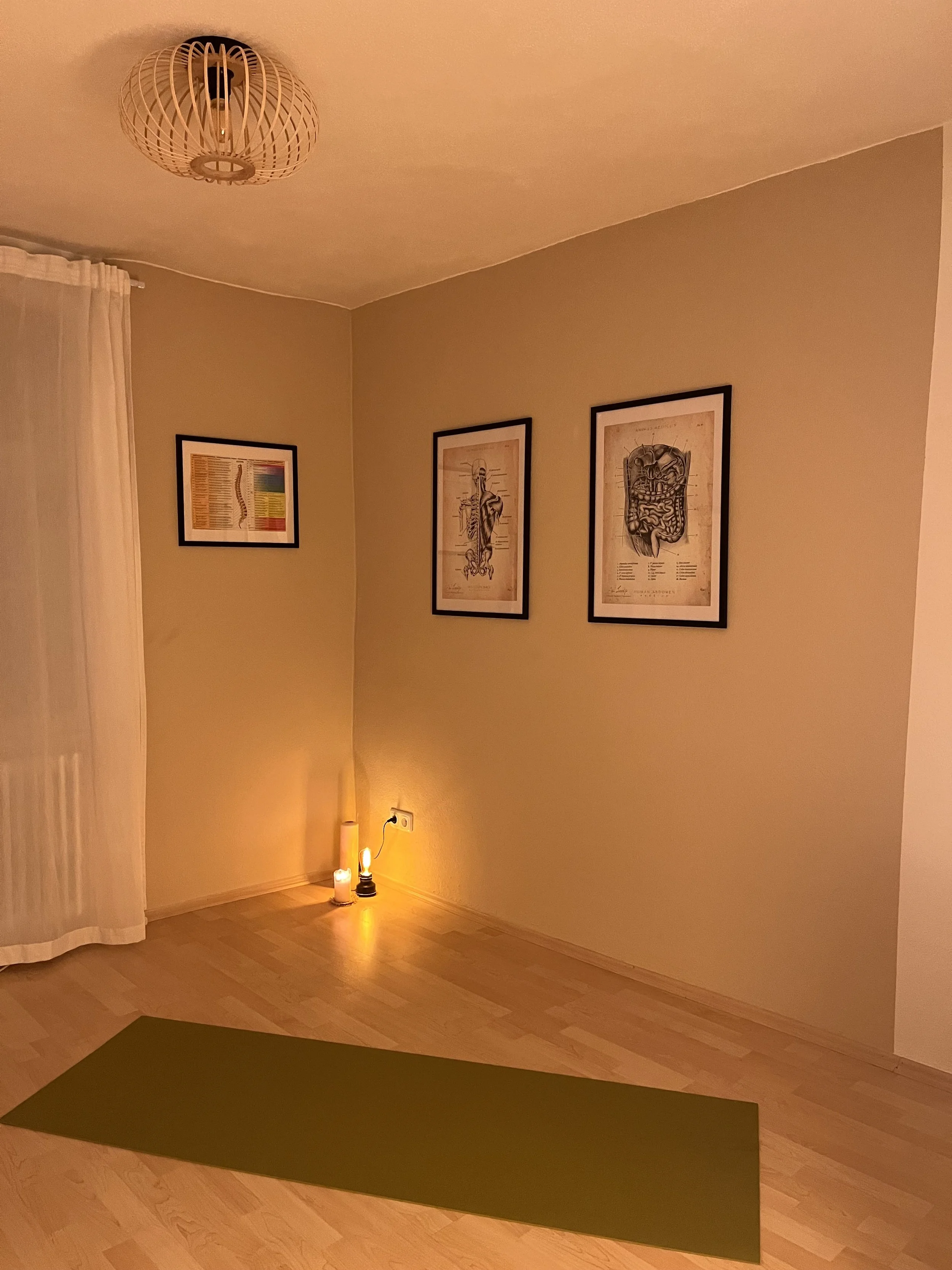 Ein Raum mit Holzfußboden, einem grünen Yogamatte, einem weißen Vorhang, drei Bilder mit anatomischen Zeichnungen an der Wand, eine kleine Lichtquelle auf dem Boden, die drei Kerzen beleuchtet.
