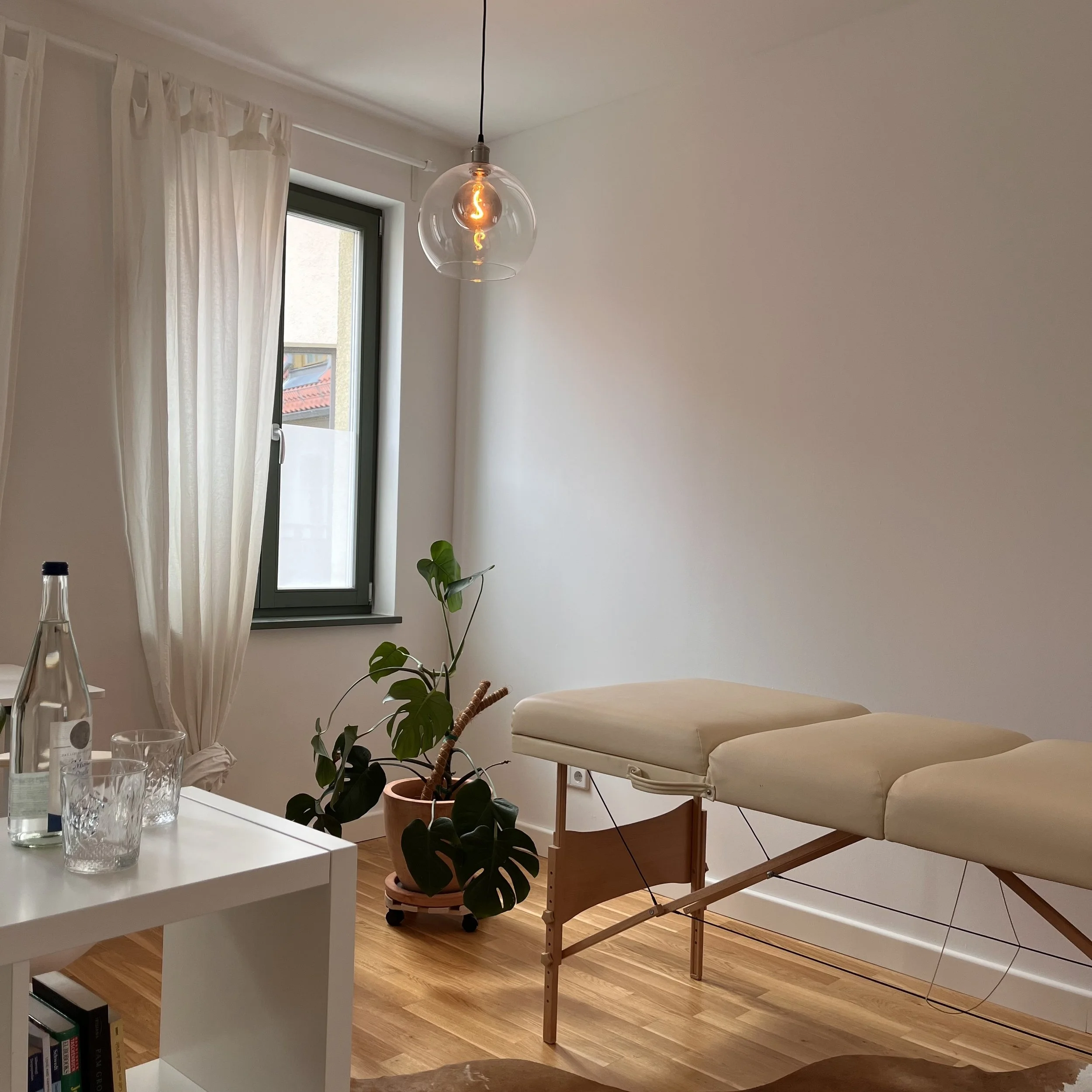 Ein Behandlungsraum mit Behandlungsliege, Pflanzen, Tisch mit Wasserflasche und Gläser, Fenster mit weißen Vorhängen, hängende Lampe.