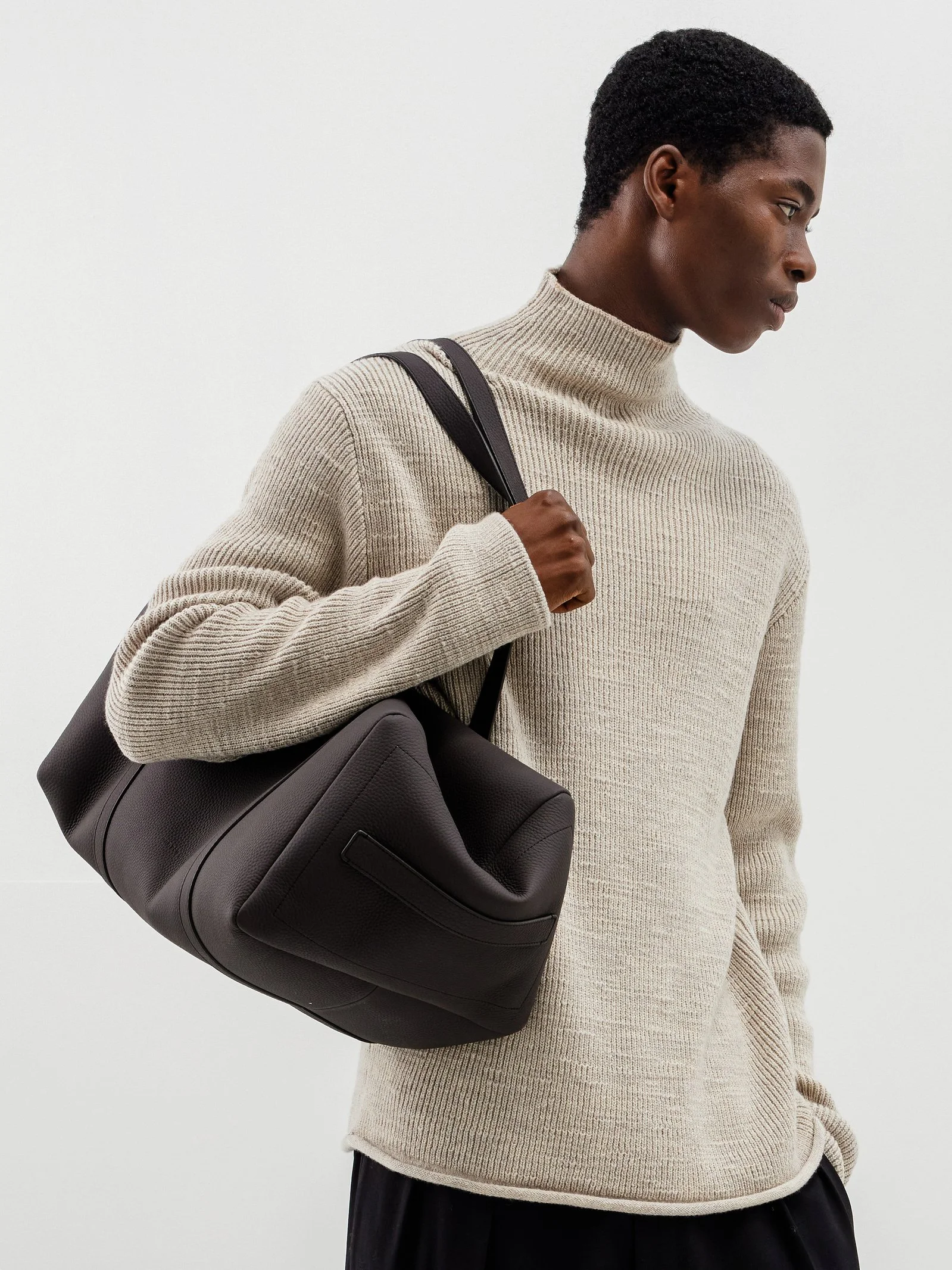 A woman with dark skin and short curly hair wearing a beige turtleneck sweater and carrying a black leather bag over her shoulder, standing against a plain white background.