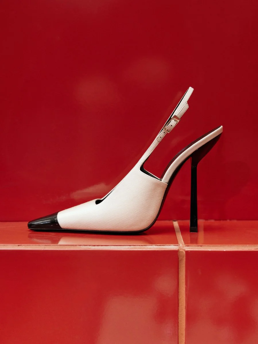 High-heeled shoe with a pointed black toe, white body, and a black stiletto heel, placed on red tiles with a red background.
