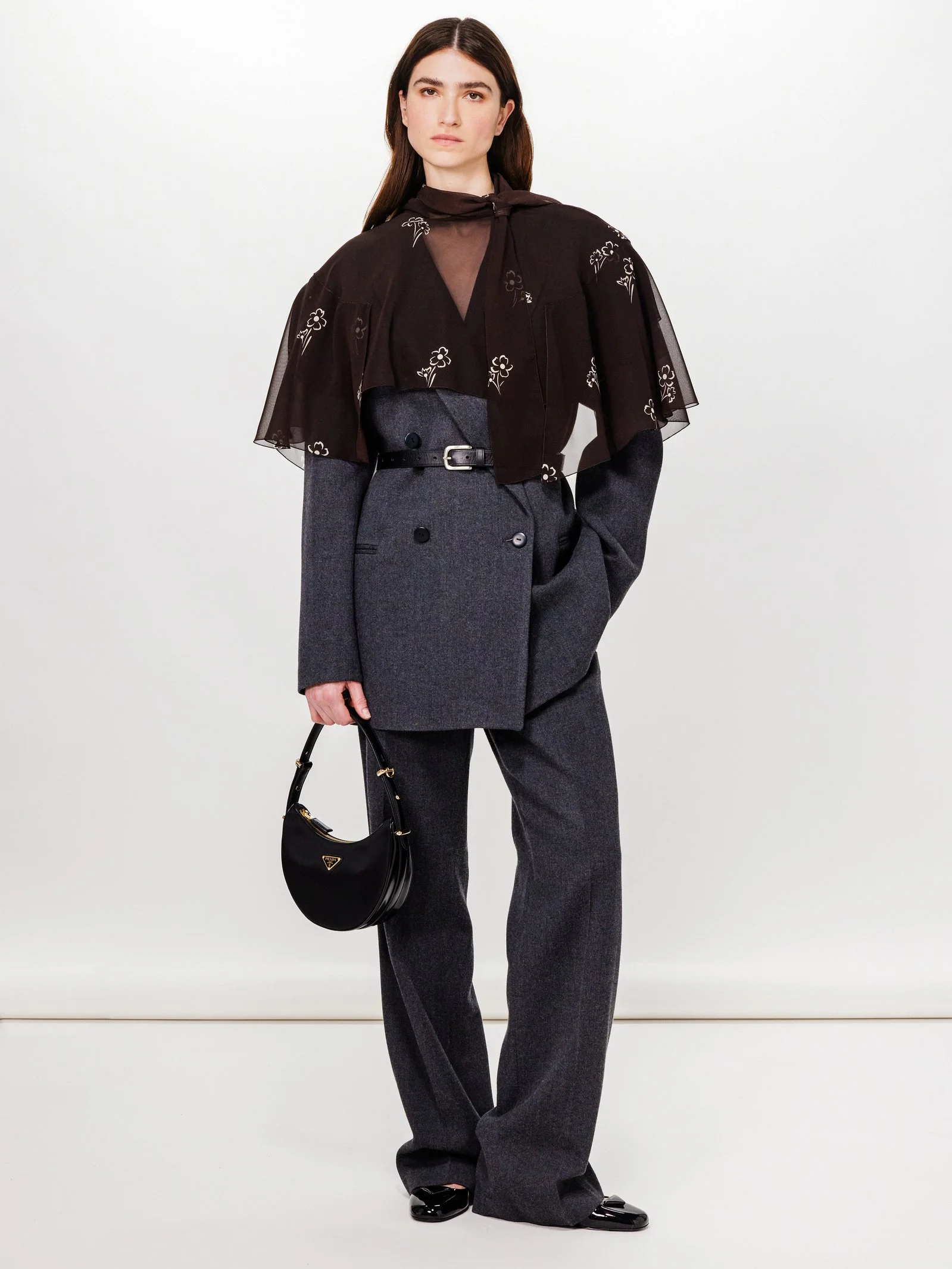 A woman with long dark hair wearing a black sheer blouse with floral embroidery over a dark blazer, paired with wide-leg gray trousers, holding a black Prada handbag, and standing against a neutral background.