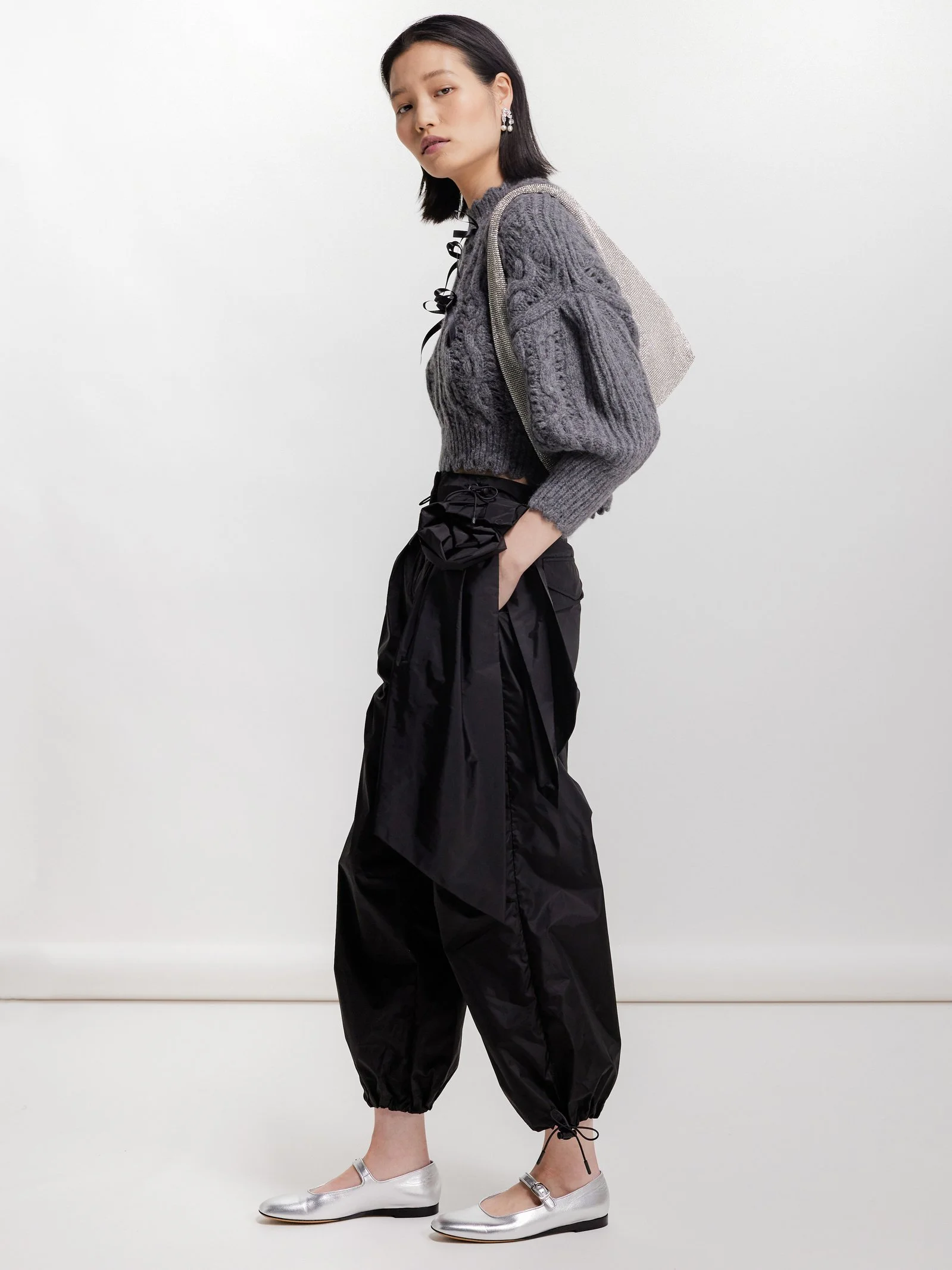 A young woman with shoulder-length black hair wearing a gray cable-knit sweater, black loose-fitting pants, and silver flats. She has earrings and carries a gray backpack over her shoulder. She is standing against a plain white background.