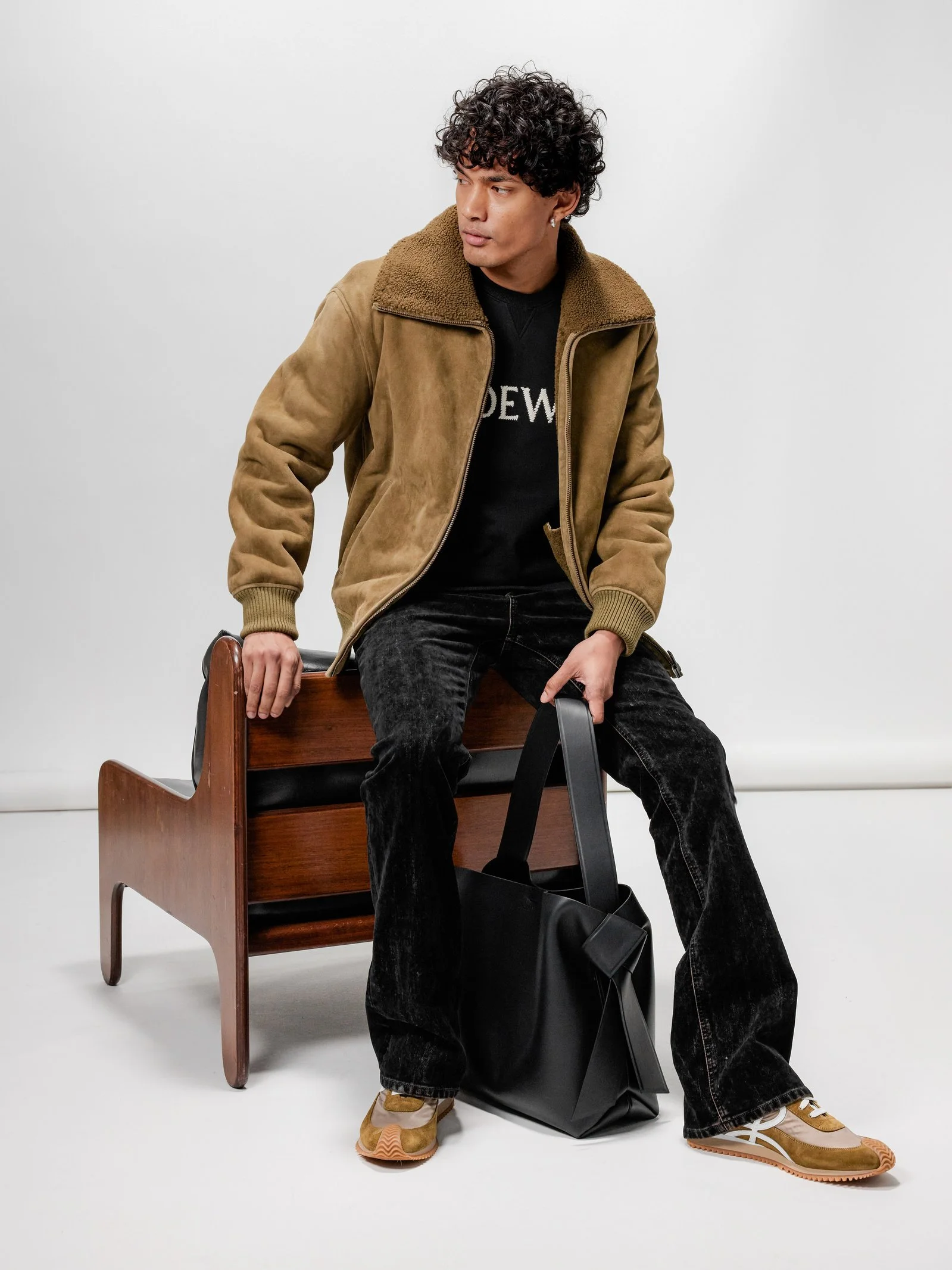 A young man with curly hair sitting on a wooden bench, wearing a brown jacket, black pants, and sneakers, holding a black bag.