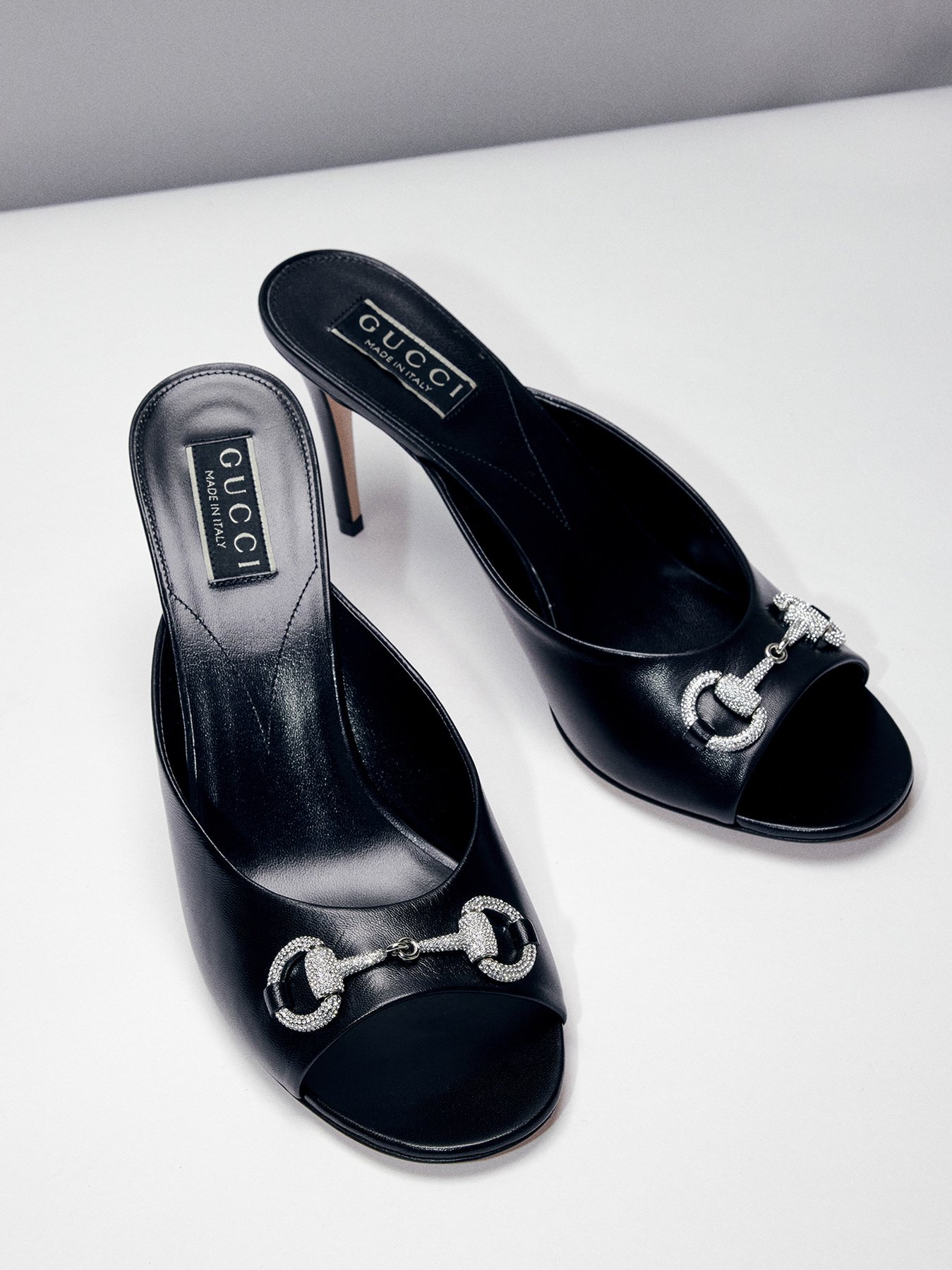Black Gucci high-heeled mule shoes with rhinestone embellishments on the toe, displayed on a white surface.