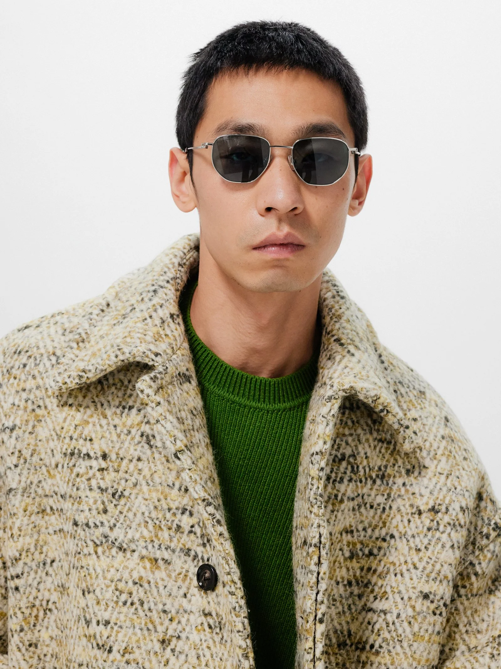 A young man with short black hair wearing sunglasses, a green sweater, and a beige patterned coat against a plain background.