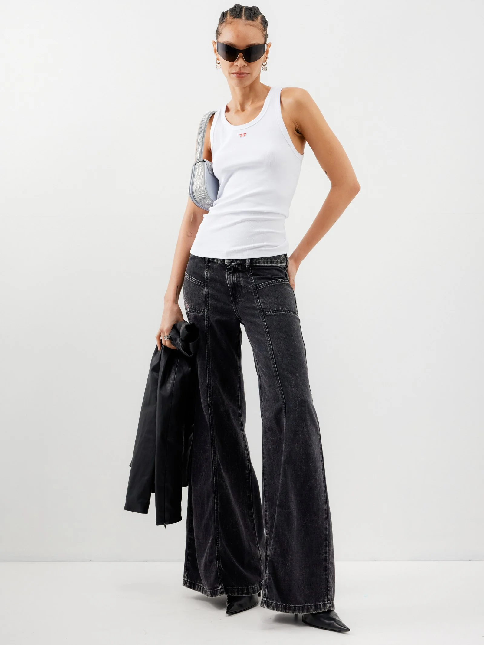 A woman standing against a plain white background, wearing large black sunglasses, a white sleeveless tank top, wide-leg black jeans, and black pointed shoes. She is holding a black jacket and a small bag with her left hand, and has her right hand in