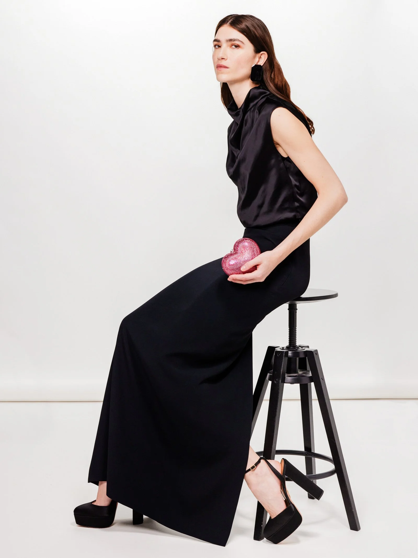 A woman with brown hair and fair skin dressed in a sleeveless black satin top and wide-leg black pants, sitting sideways on a black stool, holding a pink, glittery heart-shaped clutch, against a plain white background.