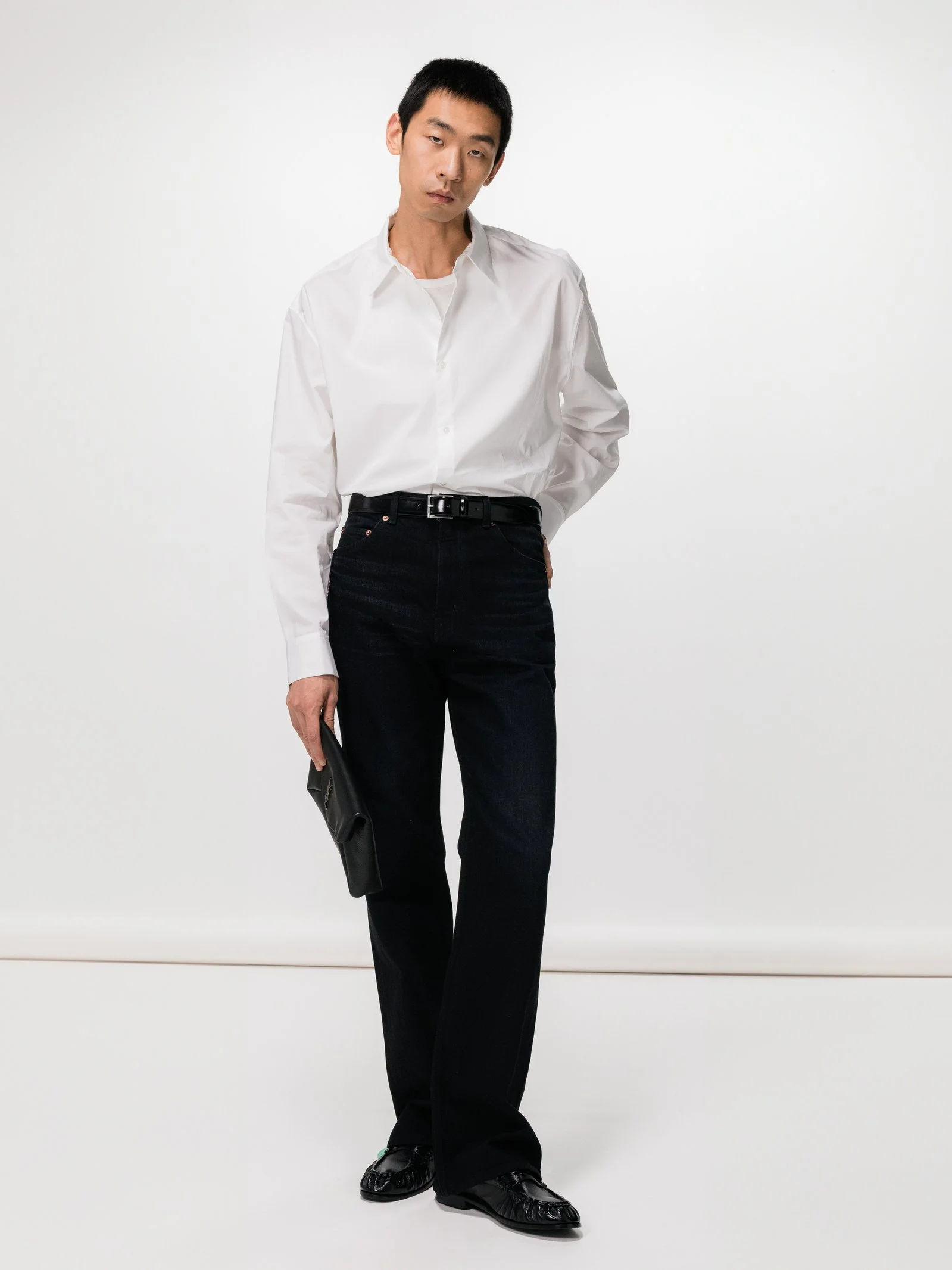 A young man with short black hair wearing a white button-up shirt, black jeans, black shoes, holding a black clutch, standing against a plain white background.
