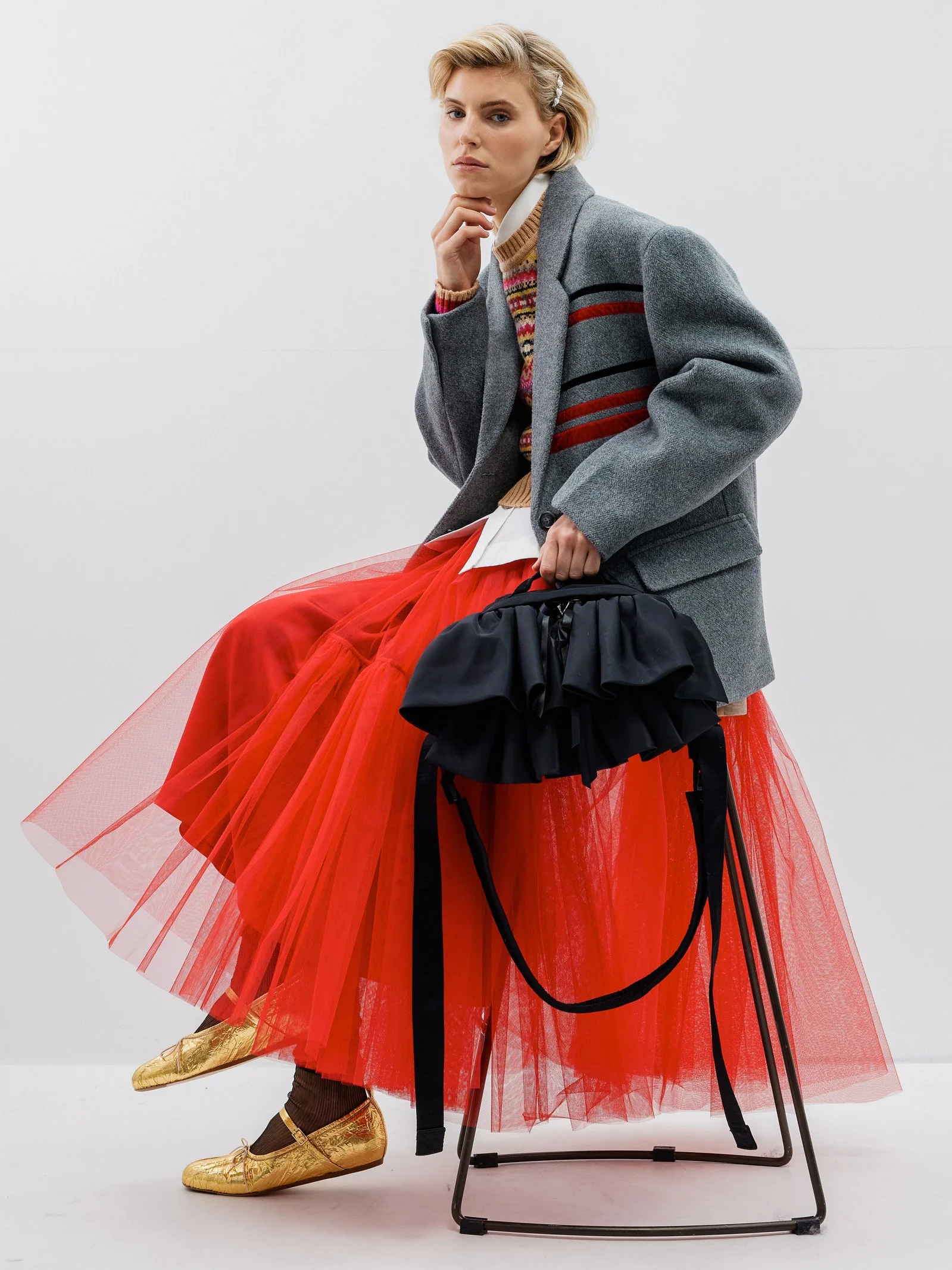 A woman sits on a chair wearing a gray blazer, colorful sweater, white shirt, red tulle skirt, gold shoes, and holds a black bag.