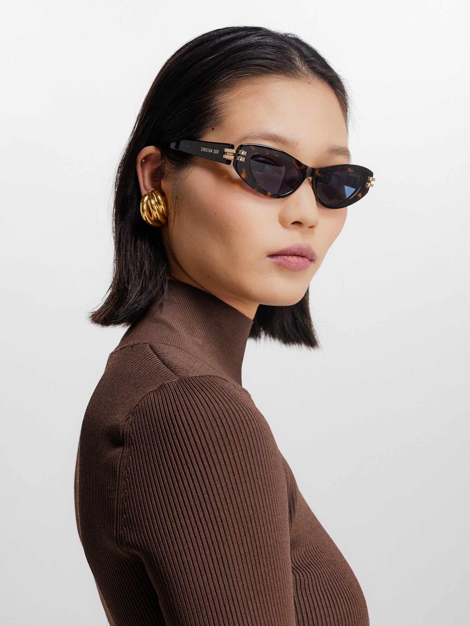 A woman with short black hair wearing black cat-eye sunglasses, gold earrings, and a brown ribbed turtleneck, looking to the side against a plain white background.