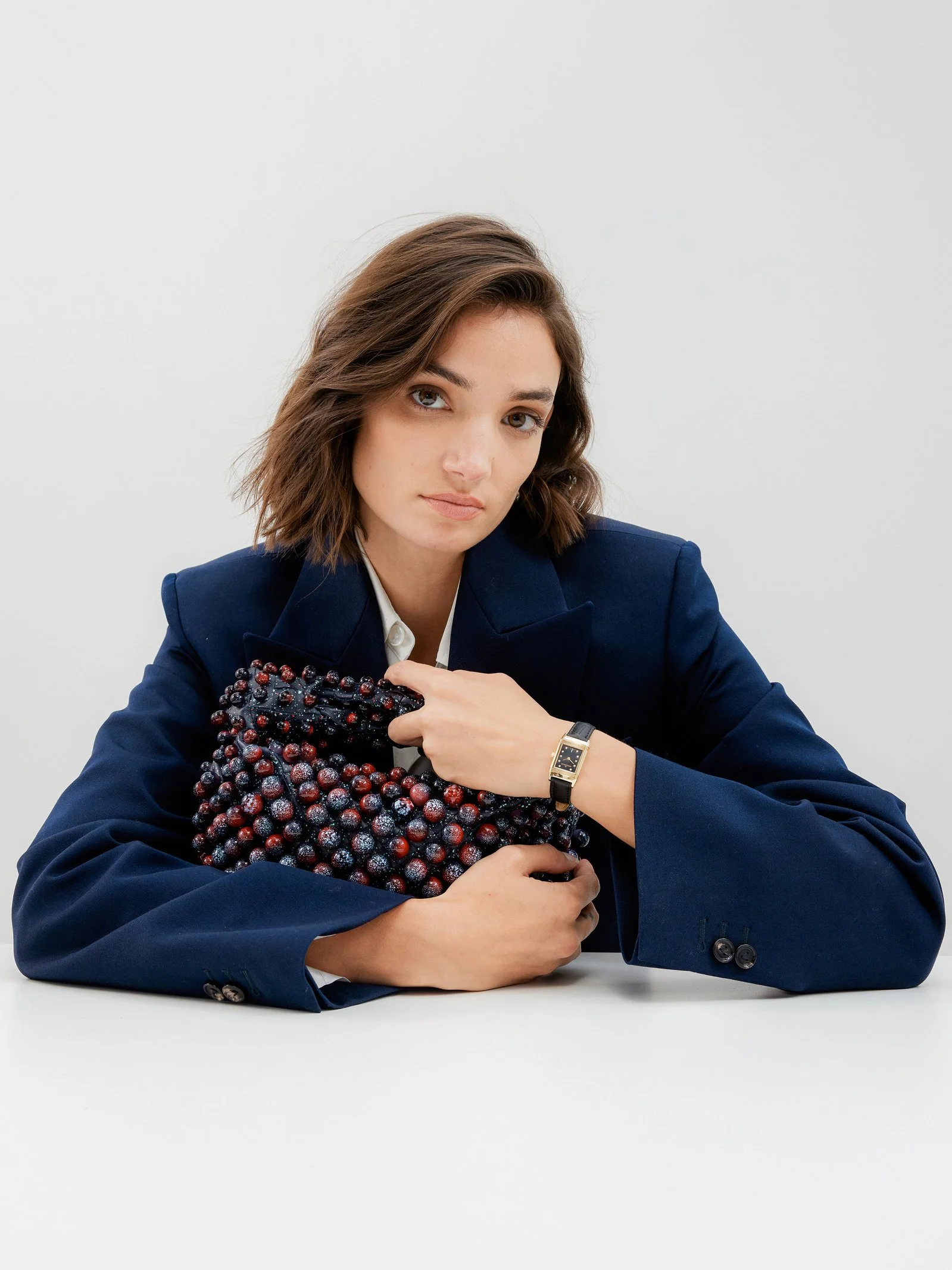 A woman with shoulder-length brown hair wearing a navy blazer, seated at a white table, holding a large, textured black and red berry-like clutch.