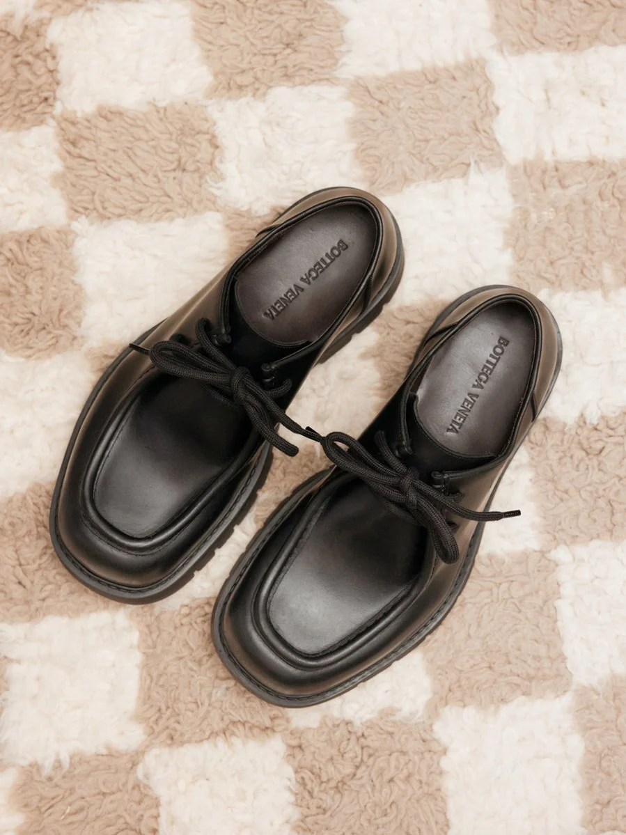 Pair of black leather designer loafers with lace-up closure on a beige and white checkered carpet.