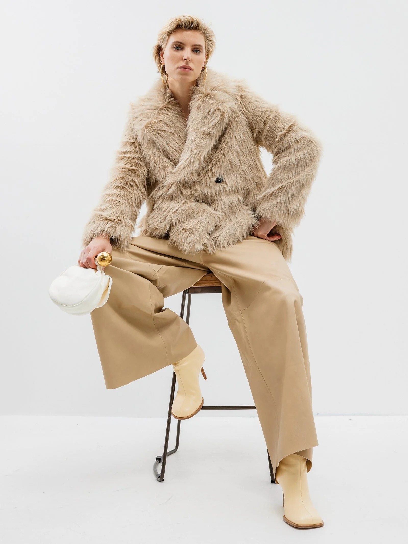 Woman sitting on a stool wearing a beige faux fur coat, wide-leg beige pants, and matching beige boots, holding a white clutch with a gold clasp.