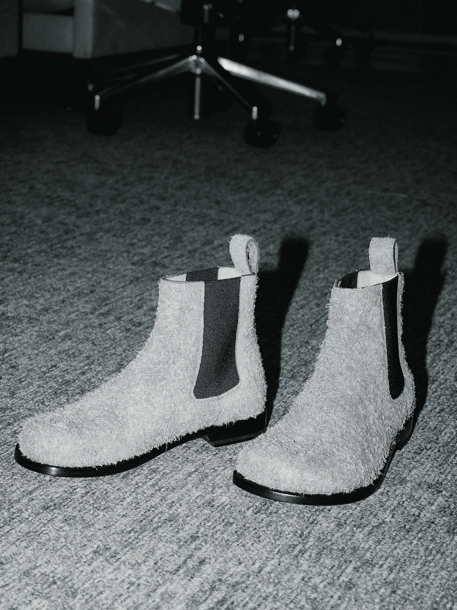 Pair of light-colored suede ankle boots with black elastic side panels on a gray carpeted floor, with a rolling chair in the background.