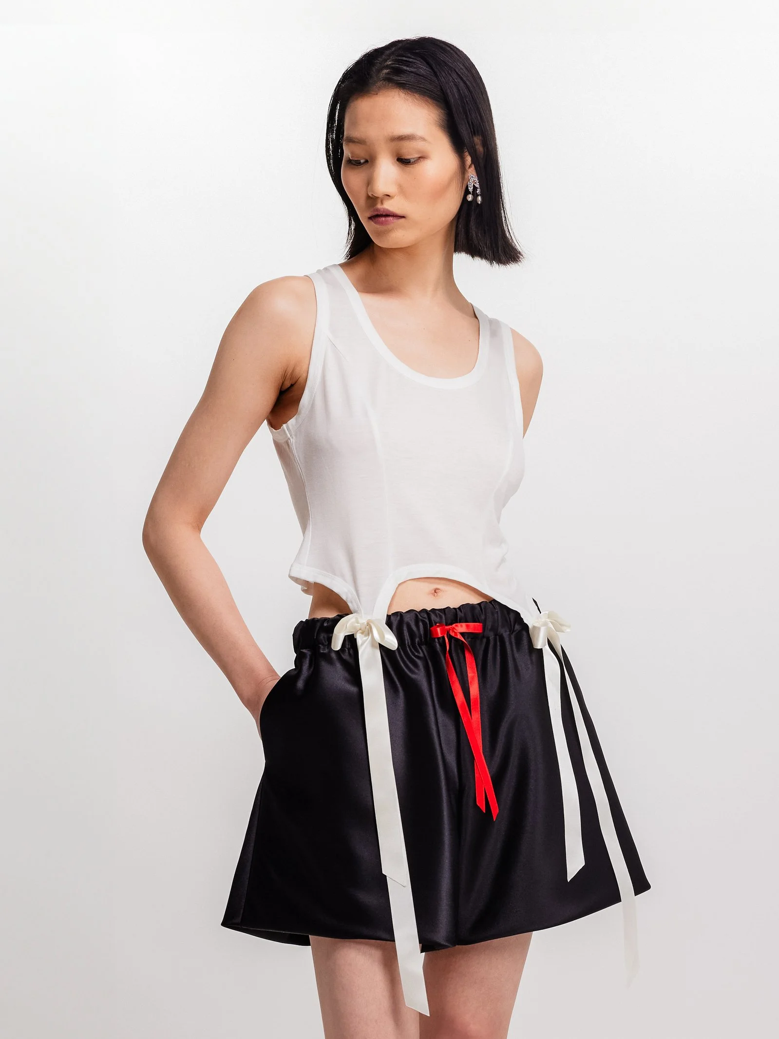 Young woman with short black hair wearing a white sleeveless crop top and black shorts with white and red ribbons, standing against a white background.