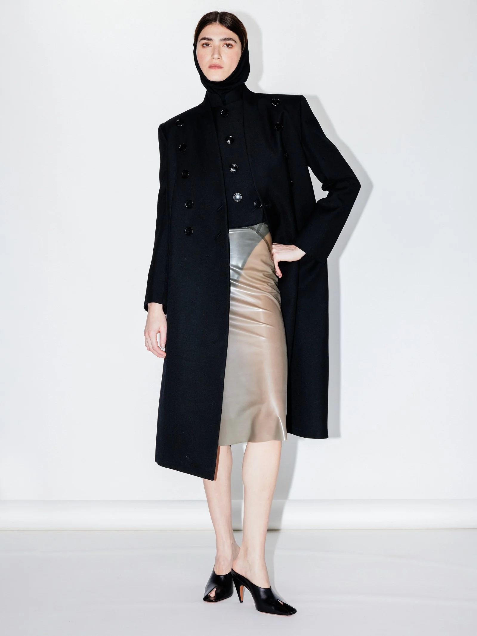A woman wearing a black coat, beige leather skirt, black high heels, and a black headscarf poses against a white background.