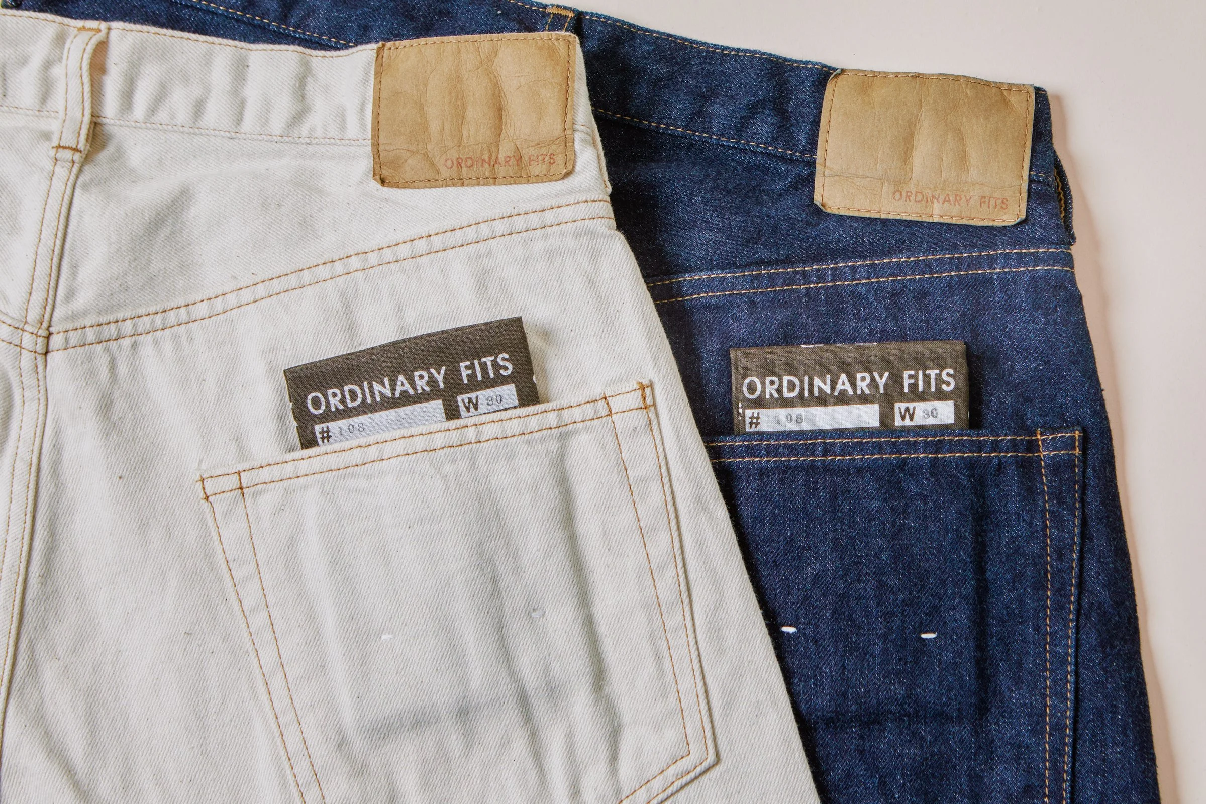 Close-up of the back pockets of two pairs of jeans, one white and one dark blue, with visible brown leather brand tags and tags that say 'ORDINARY FITS.'