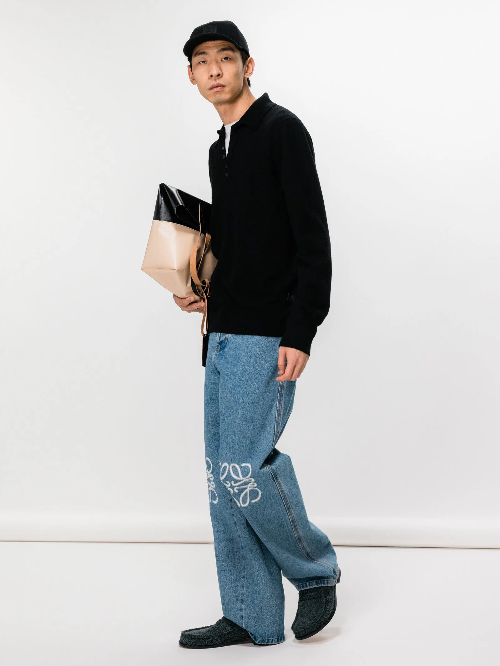 A young man in a black cricket cap, black long-sleeve shirt, and blue jeans with white embroidery, holding a paper shopping bag, standing against a plain white background.