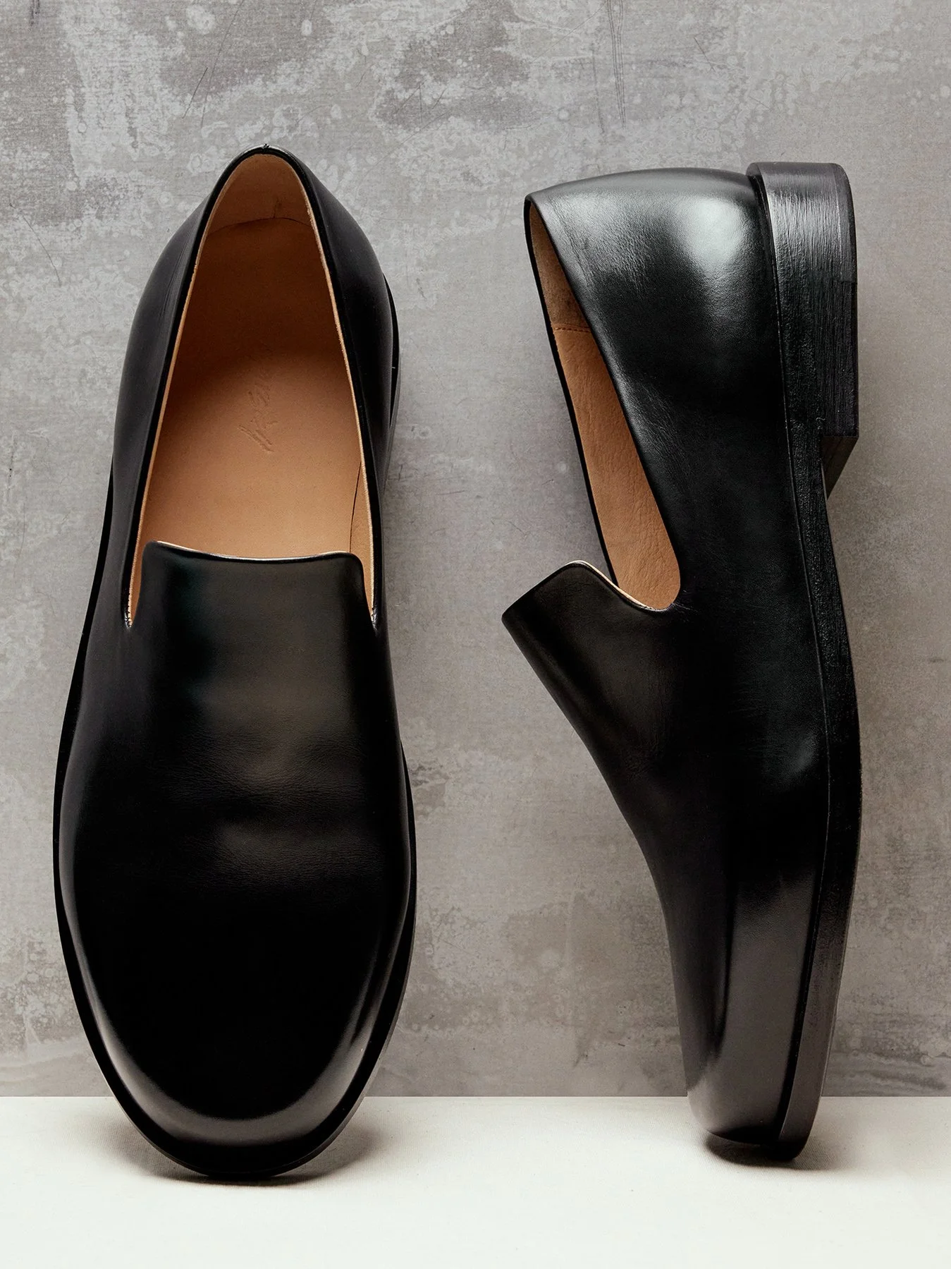 Pair of black leather dress shoes placed on a white surface against a textured gray wall, with one shoe showing the top view and the other showing the side view.