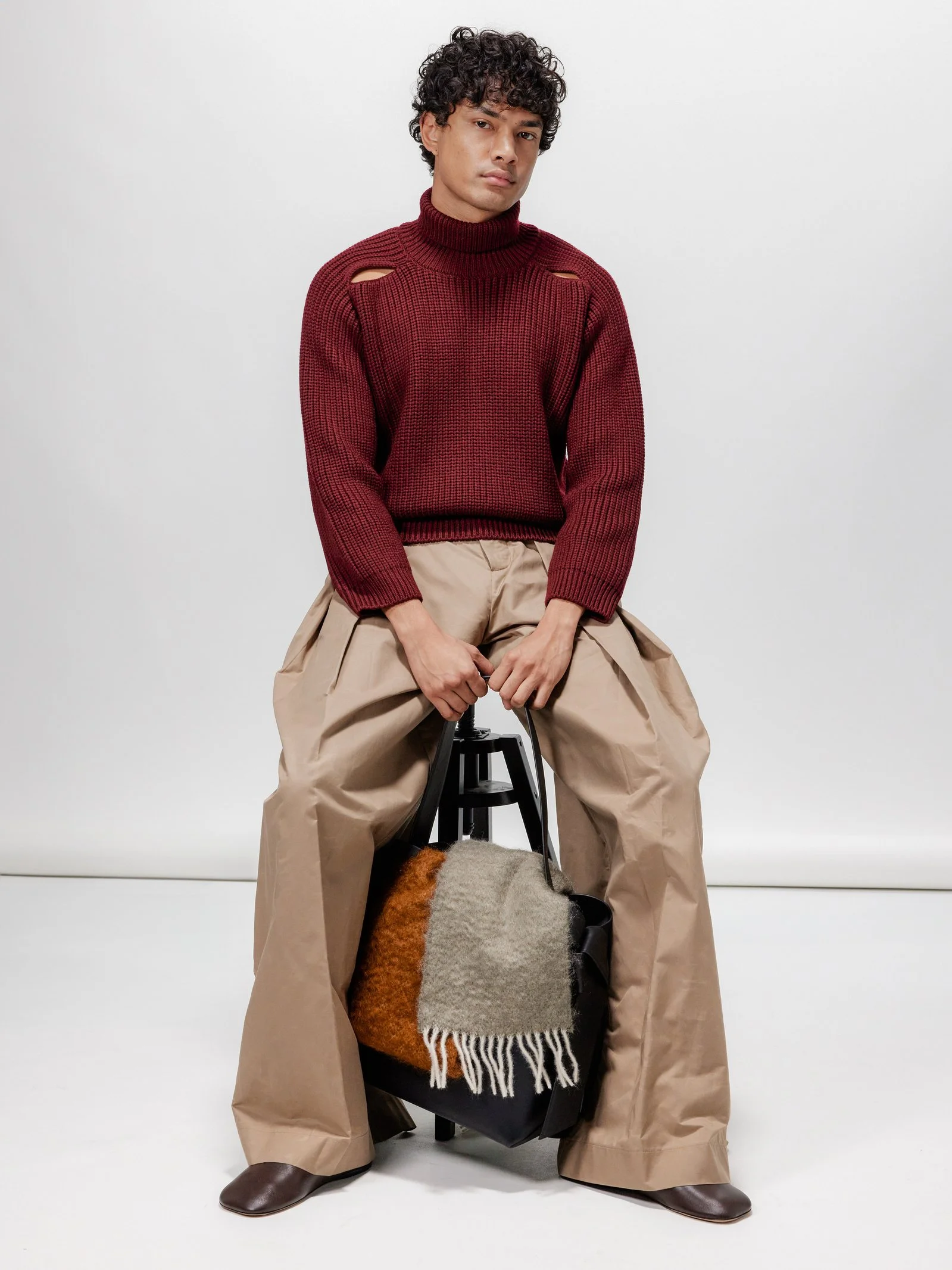 Man sitting on a stool holding a black bag with a multicolored scarf, wearing beige wide-leg pants and a red sweater with cutouts on the shoulders, in front of a plain white background.