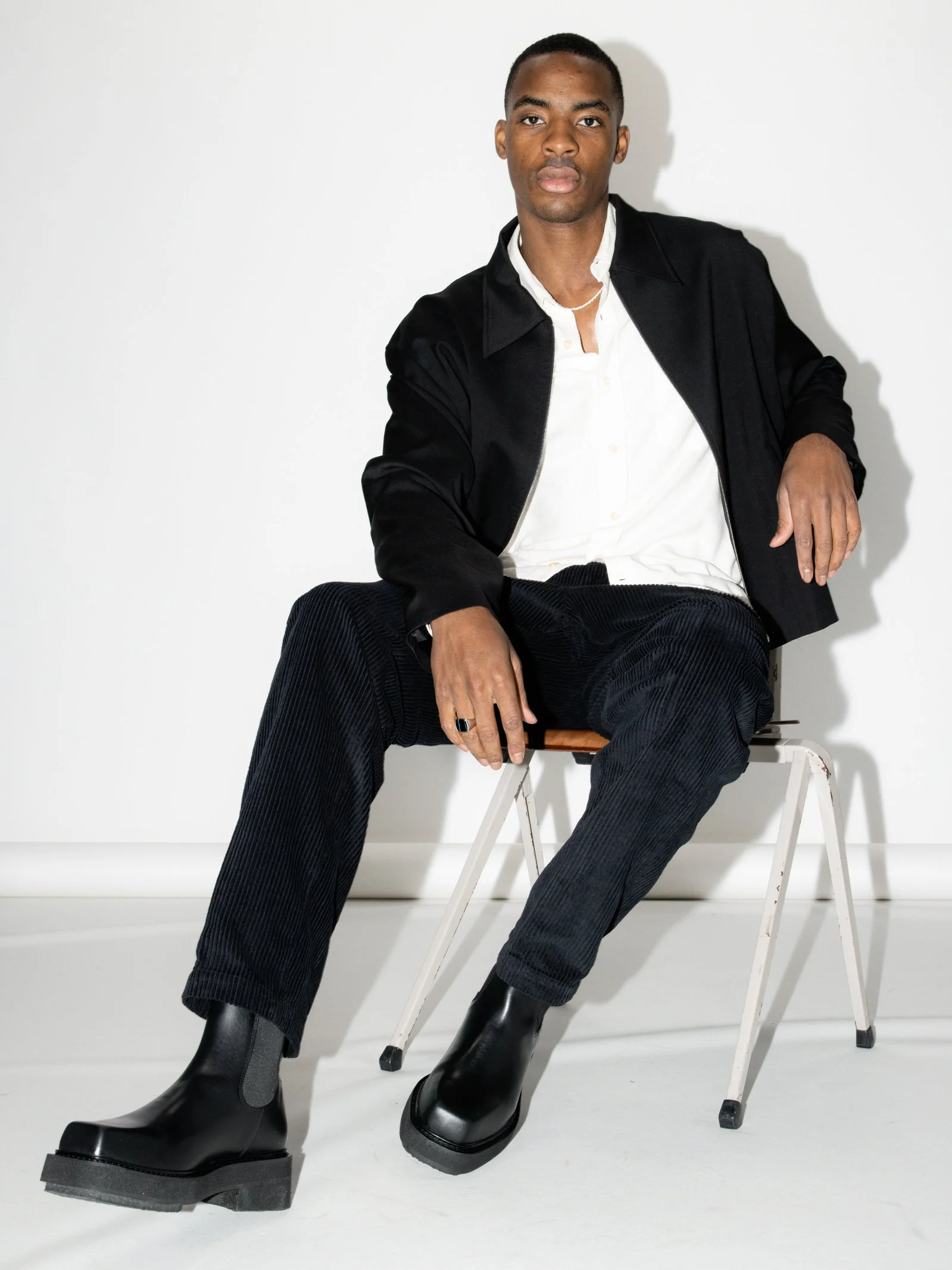 A young man wearing a black jacket, white shirt, black pants, and black boots sitting on a white chair against a plain white wall.