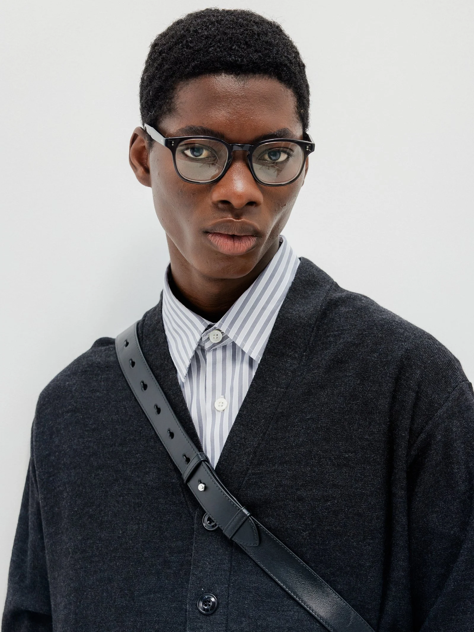 Young man with dark skin, short hair, glasses, wearing a striped button-up shirt under a dark blazer, standing against a plain white background.