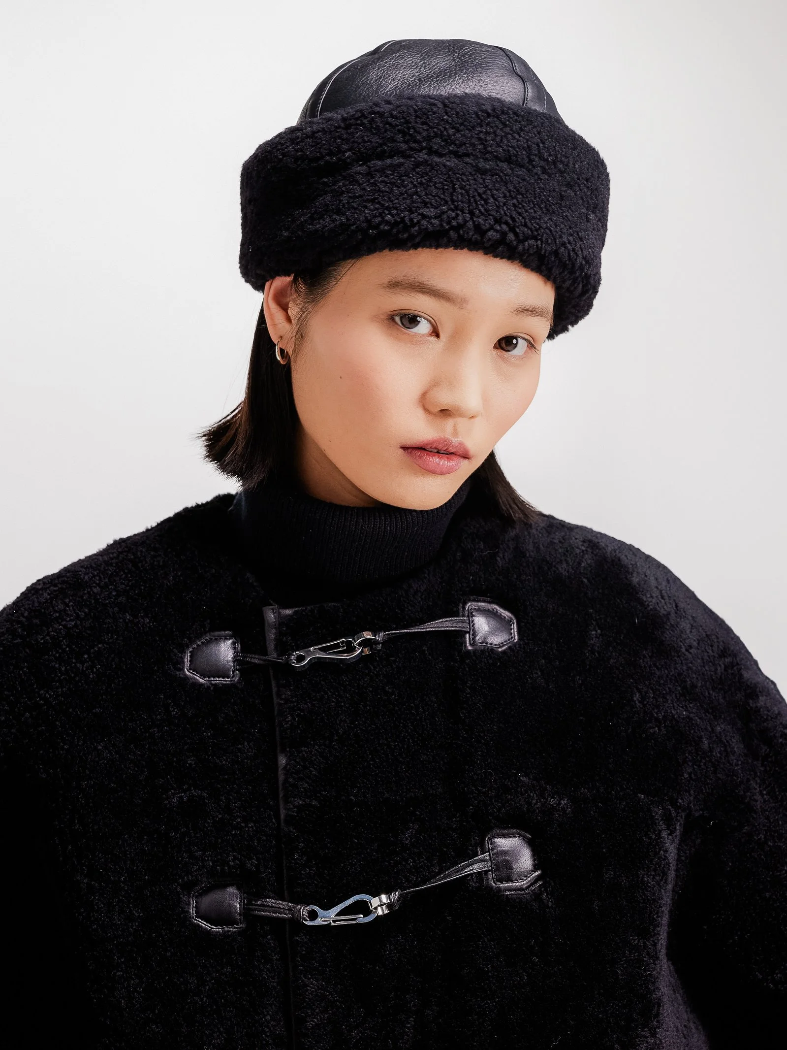 A young woman with short dark hair wearing a black fur coat, black turtleneck, and a black hat with a leather detail on top, against a plain white background.