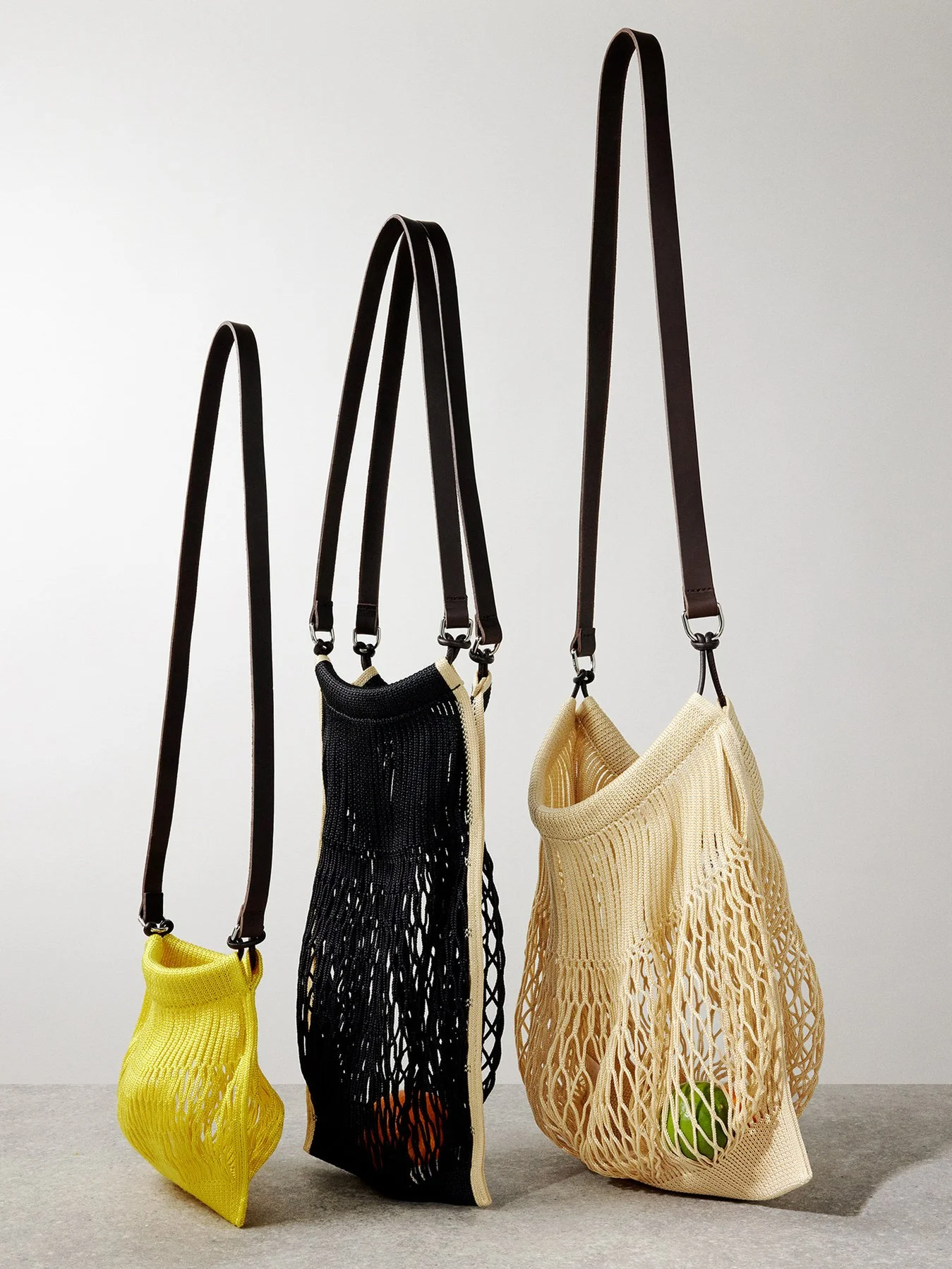 Three netted bags on display, a small yellow bag, a black bag with an orange ball inside, and a beige bag with a green ball inside, each with long shoulder straps.