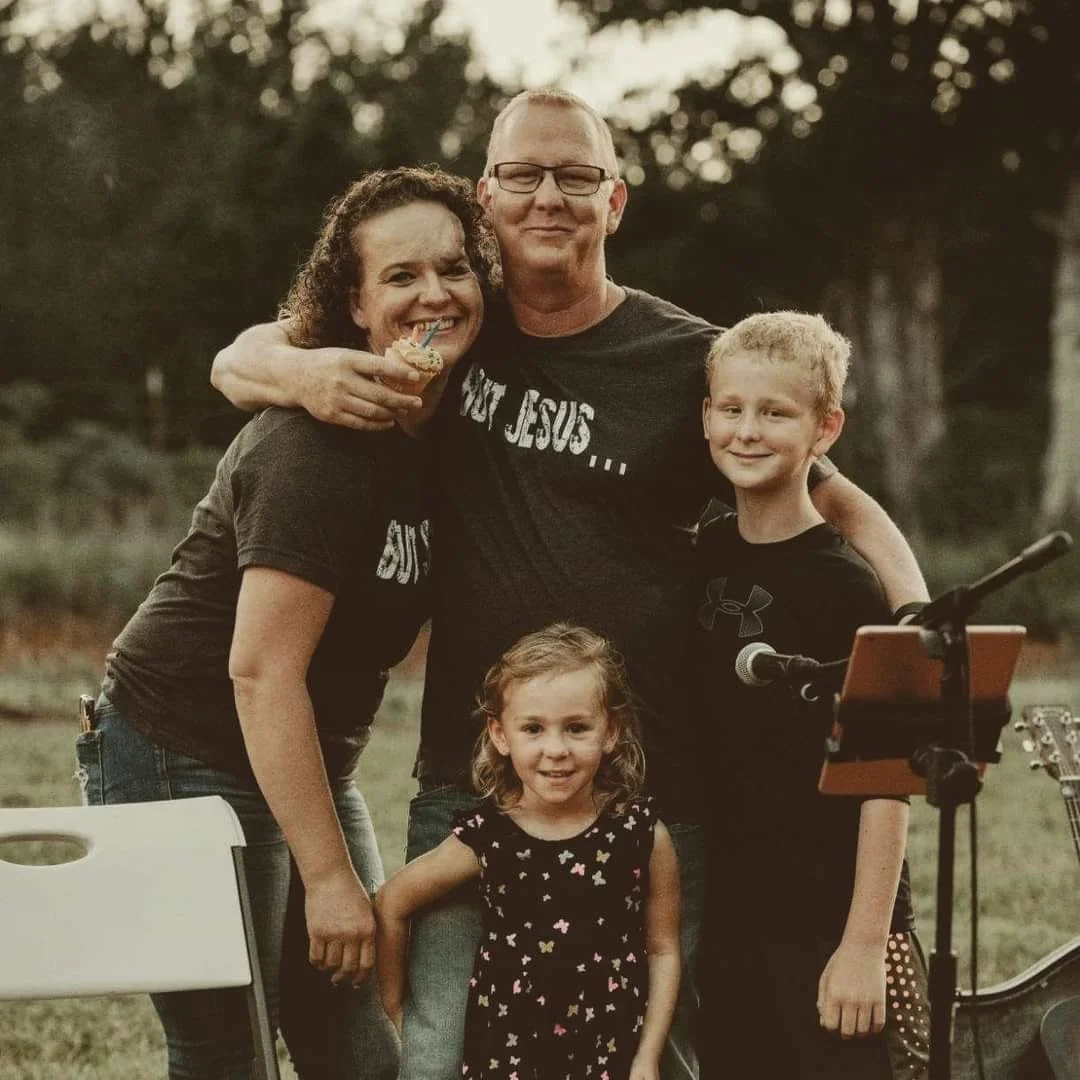 A family of five, including two adults and three children, standing outdoors during sunset. They are smiling and gathered closely, with microphones and music equipment nearby, suggesting a casual outdoor gathering or performance.