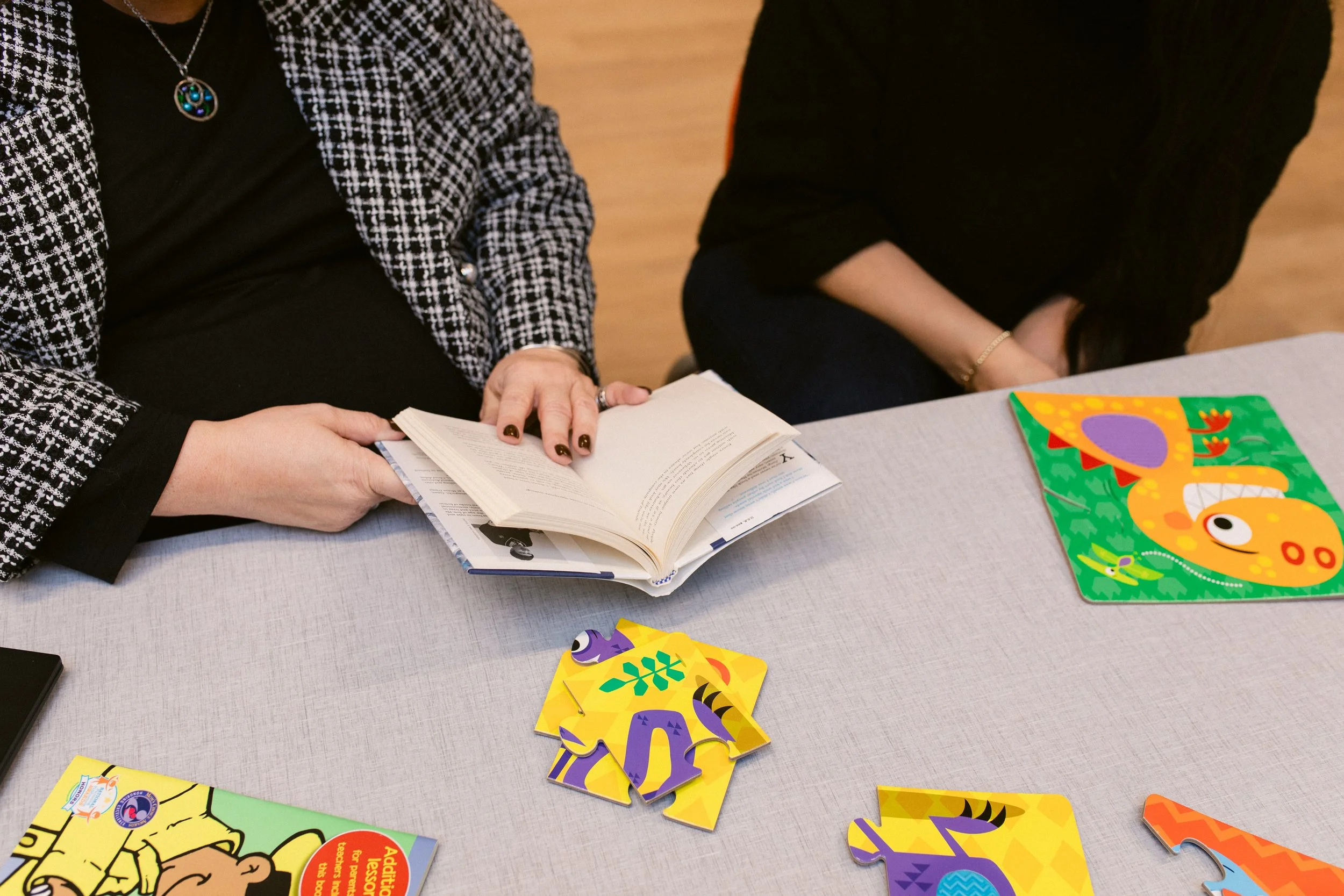 Two women sitting at a table, with one reading a book and the other resting her arms on the table. There are colorful animal-shaped puzzle pieces and a children's book with a dinosaur illustration on the table.