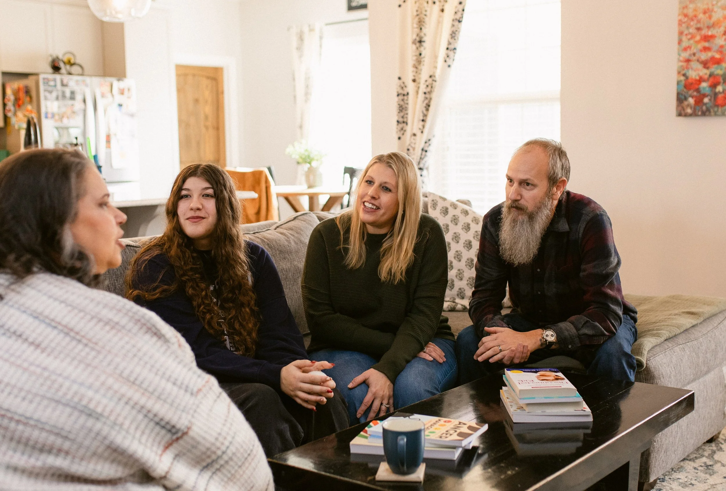 A family sitting together on a sofa in a living room, engaging in conversation with a woman, a girl, and a man with a grey beard.