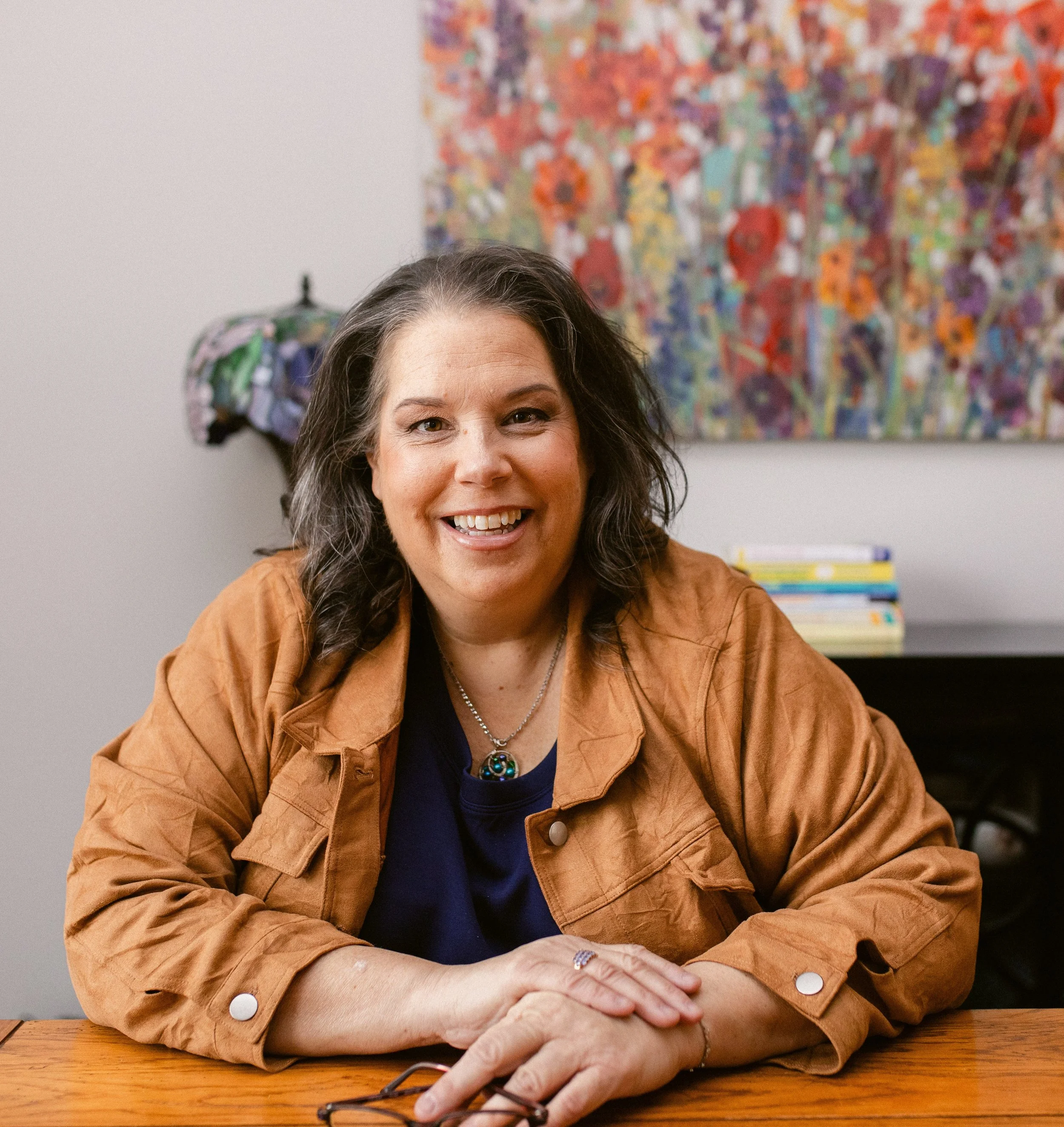 A woman with shoulder-length dark hair, smiling, wearing a tan jacket over a blue shirt, sitting at a wooden desk, with a colorful abstract painting and a stack of books in the background.
