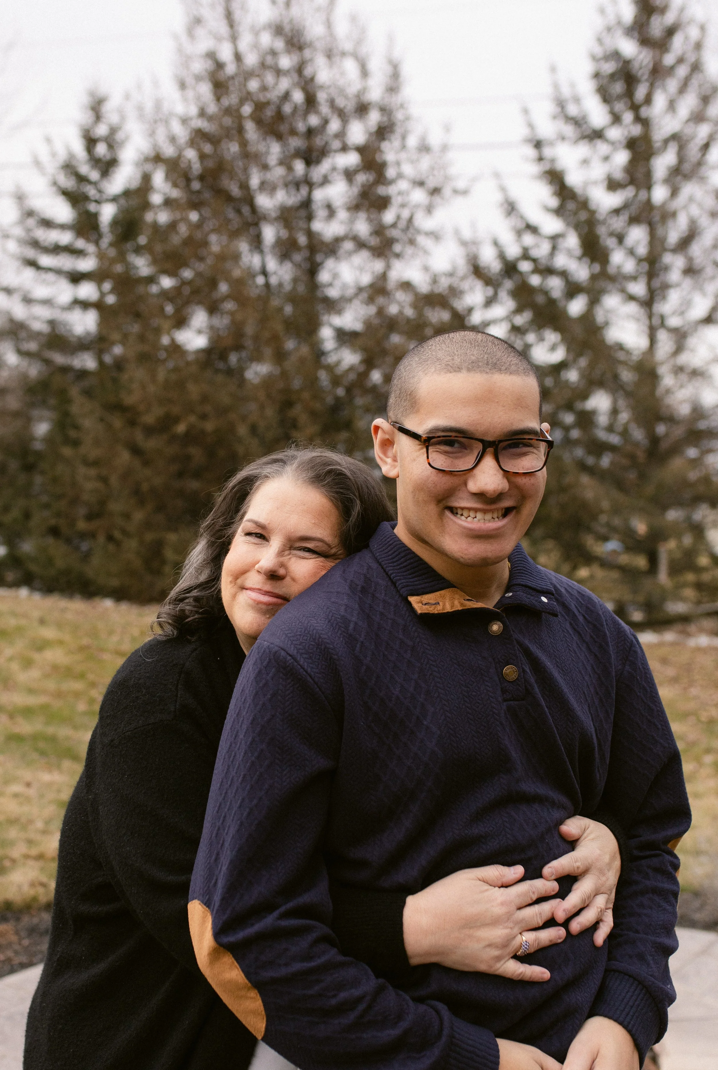 A woman stands behind a smiling young man, hugging him from behind with her hands on his stomach, outdoors with trees in the background.