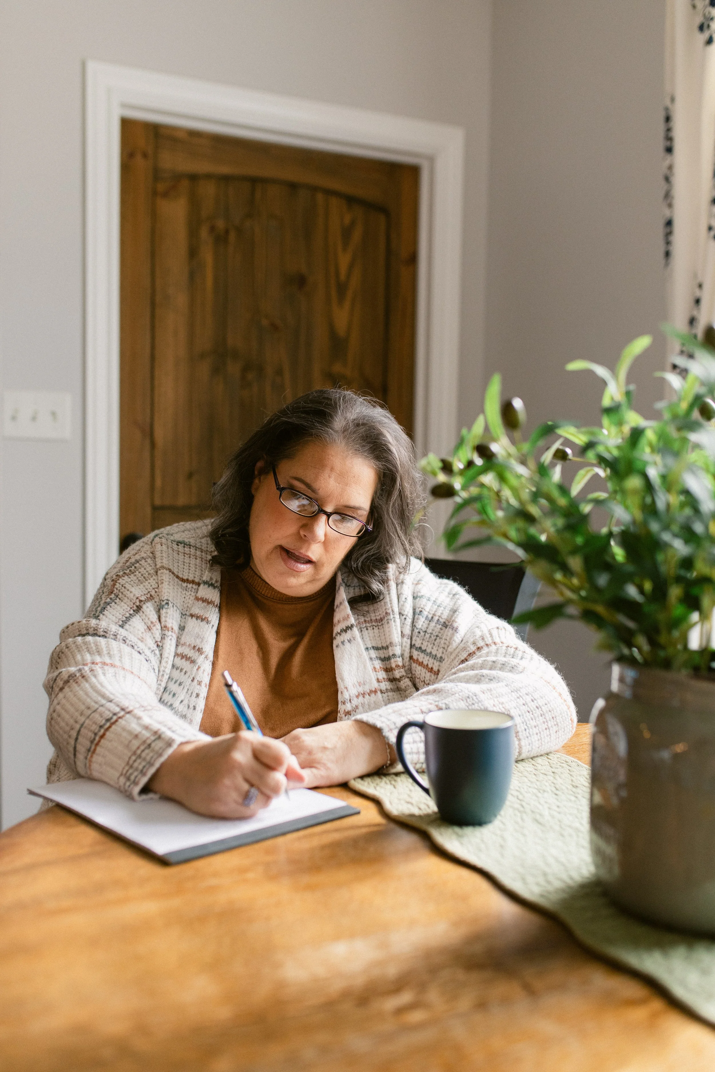 A woman with glasses and gray hair writing in a notebook at a wooden table, with a black mug and a potted plant.