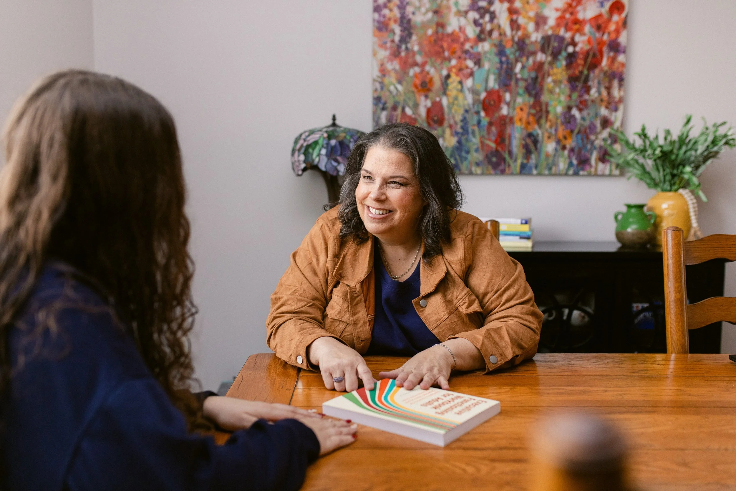 Two women sitting at a wooden table in a room. One woman is smiling, wearing a brown jacket and a navy shirt, with a book in front of her. The other woman, with long curly hair, is wearing a dark shirt and is listening. In the background, there is a colorful painting on the wall, a decorative lamp, and a table with green and yellow vases with plants.