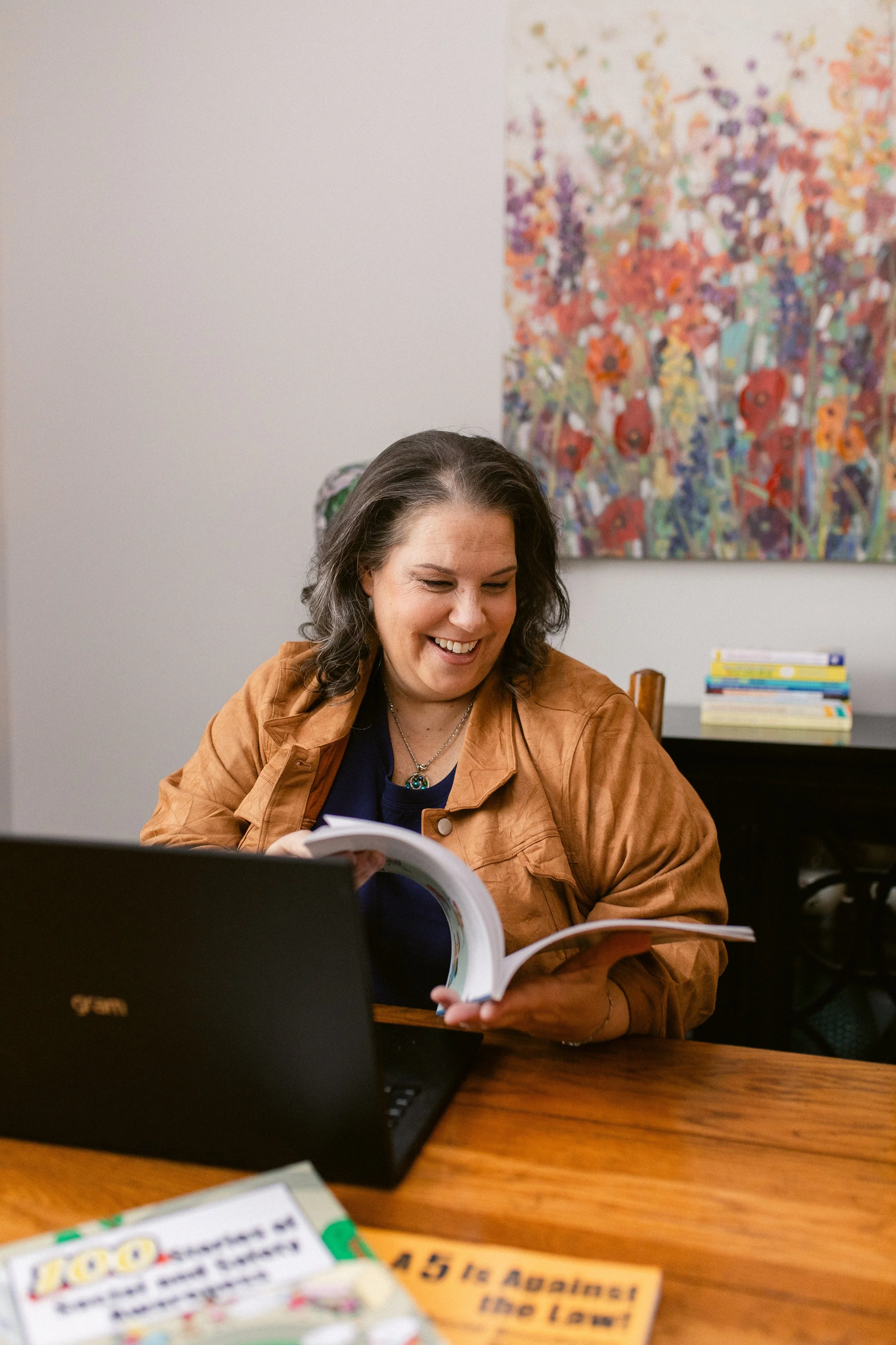 A woman smiling and looking at a book while sitting at a wooden table with a laptop open in front of her. In the background, colorful artwork hangs on the wall and there is a small stack of books on a shelf.