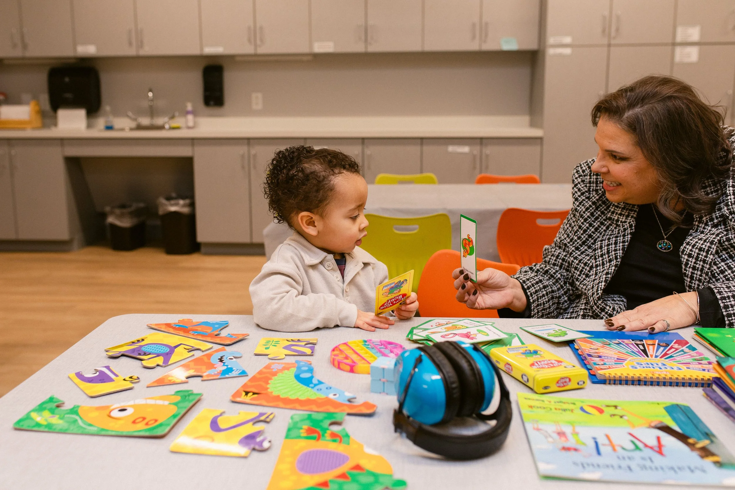 A woman and a young boy sitting at a table playing a card game with colorful animal and alphabet-themed cards in a classroom or activity room.