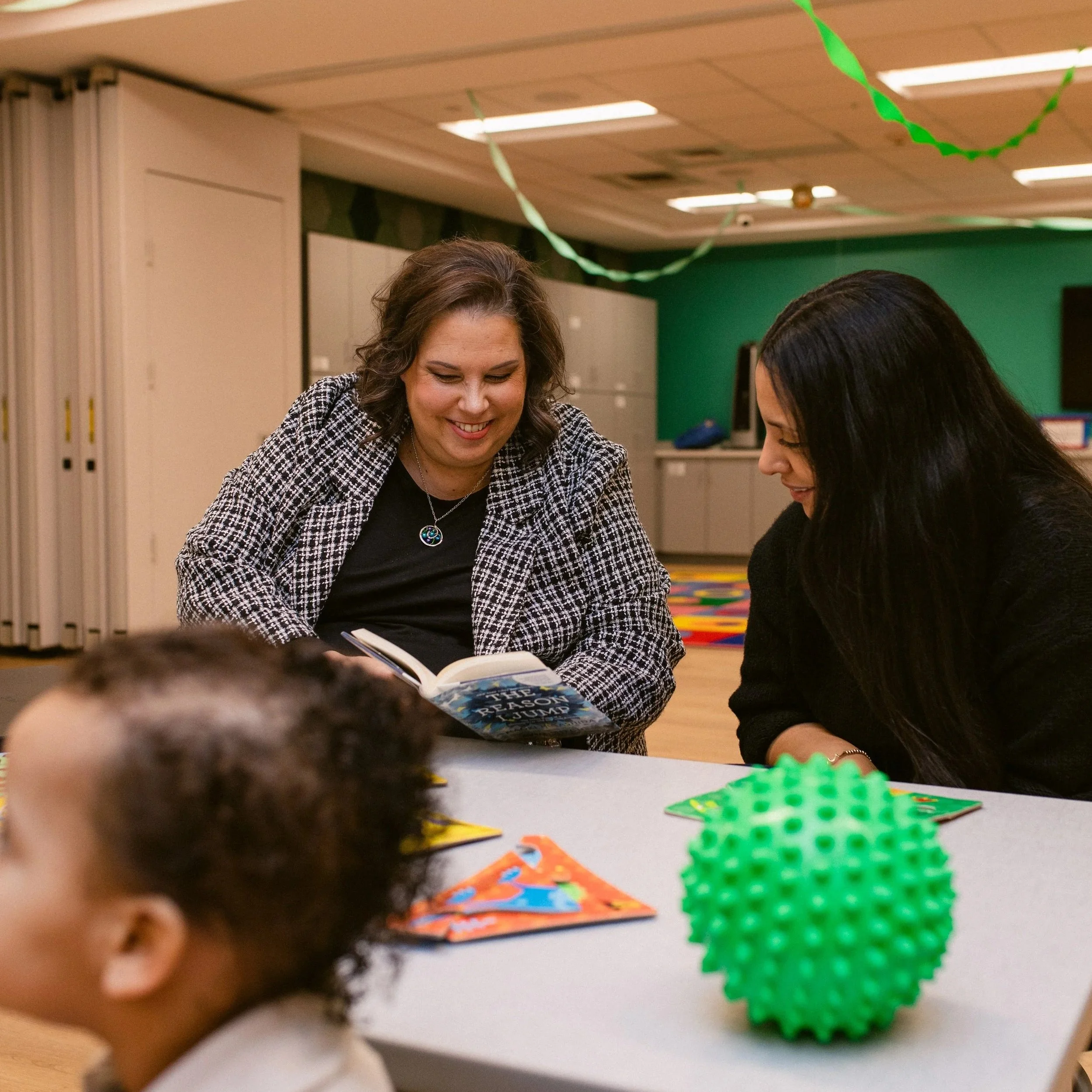 Two women reading a book together at a table in a classroom or activity room, with colorful decorations and a green wall in the background, and children in the foreground.