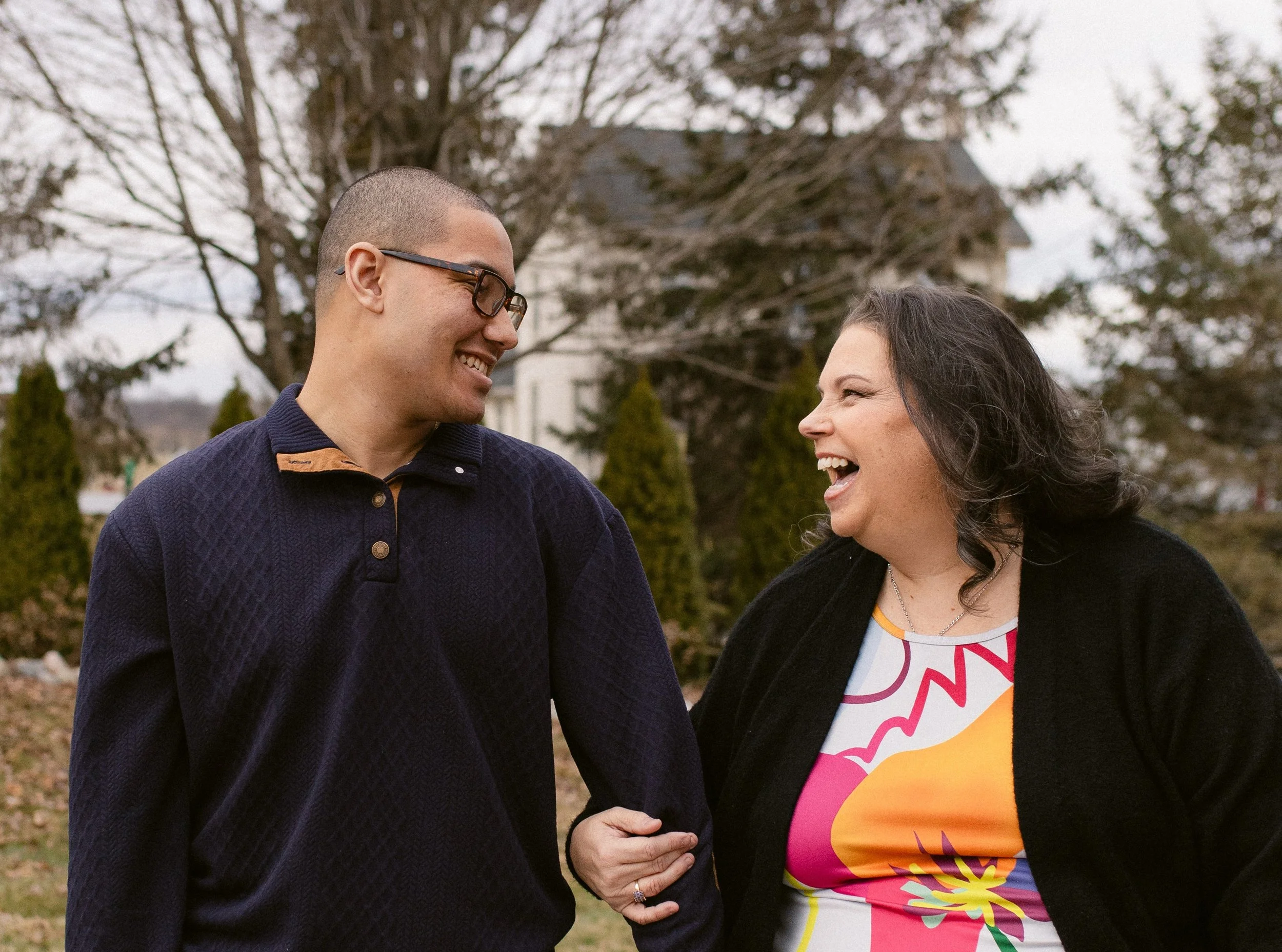 A man and a woman are laughing and smiling at each other outdoors in a park or garden, with trees and a house in the background.