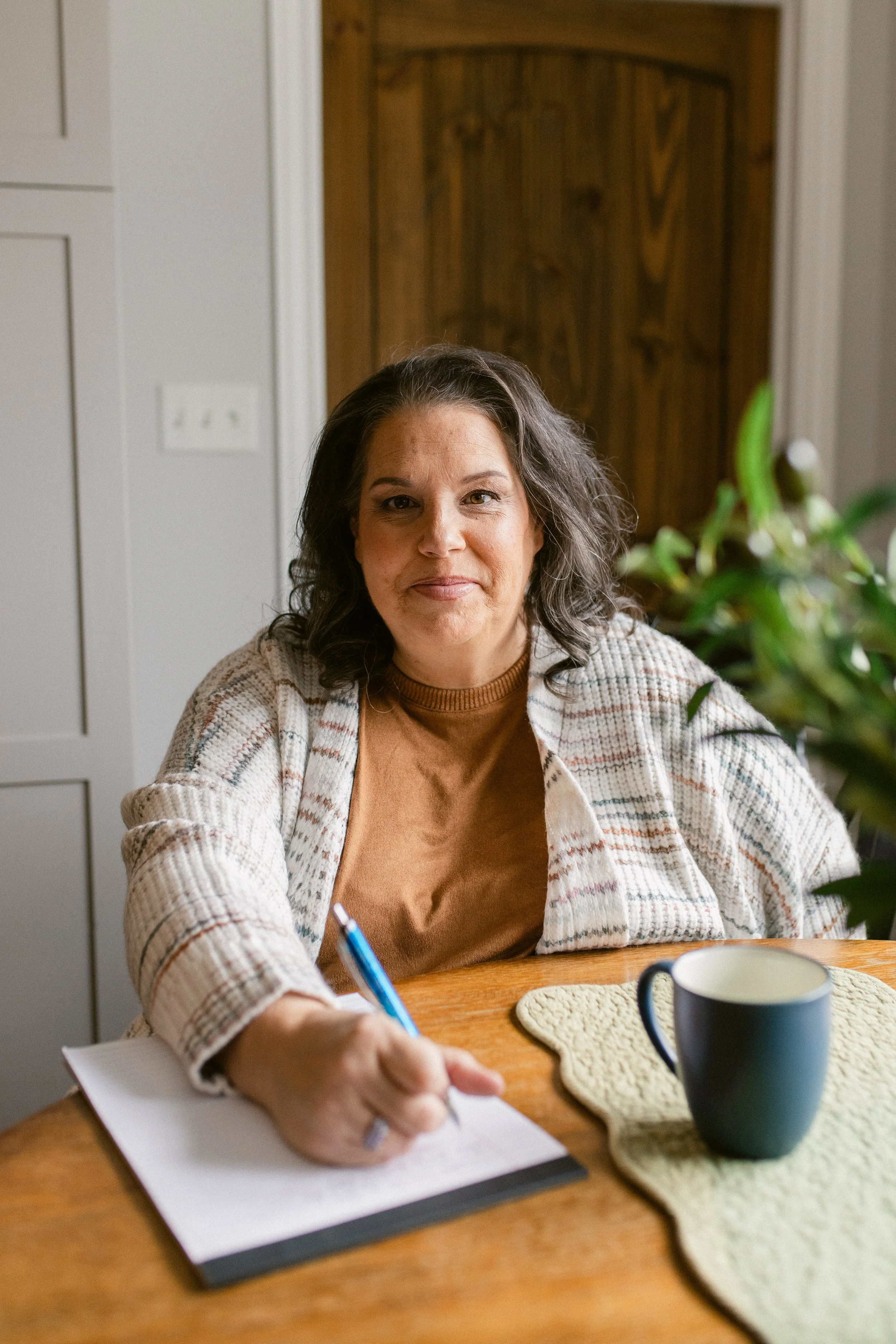 A woman with shoulder-length gray hair sitting at a wooden table, writing in a notebook with a blue pen, with a black mug and a plant on the table, in a cozy kitchen setting.