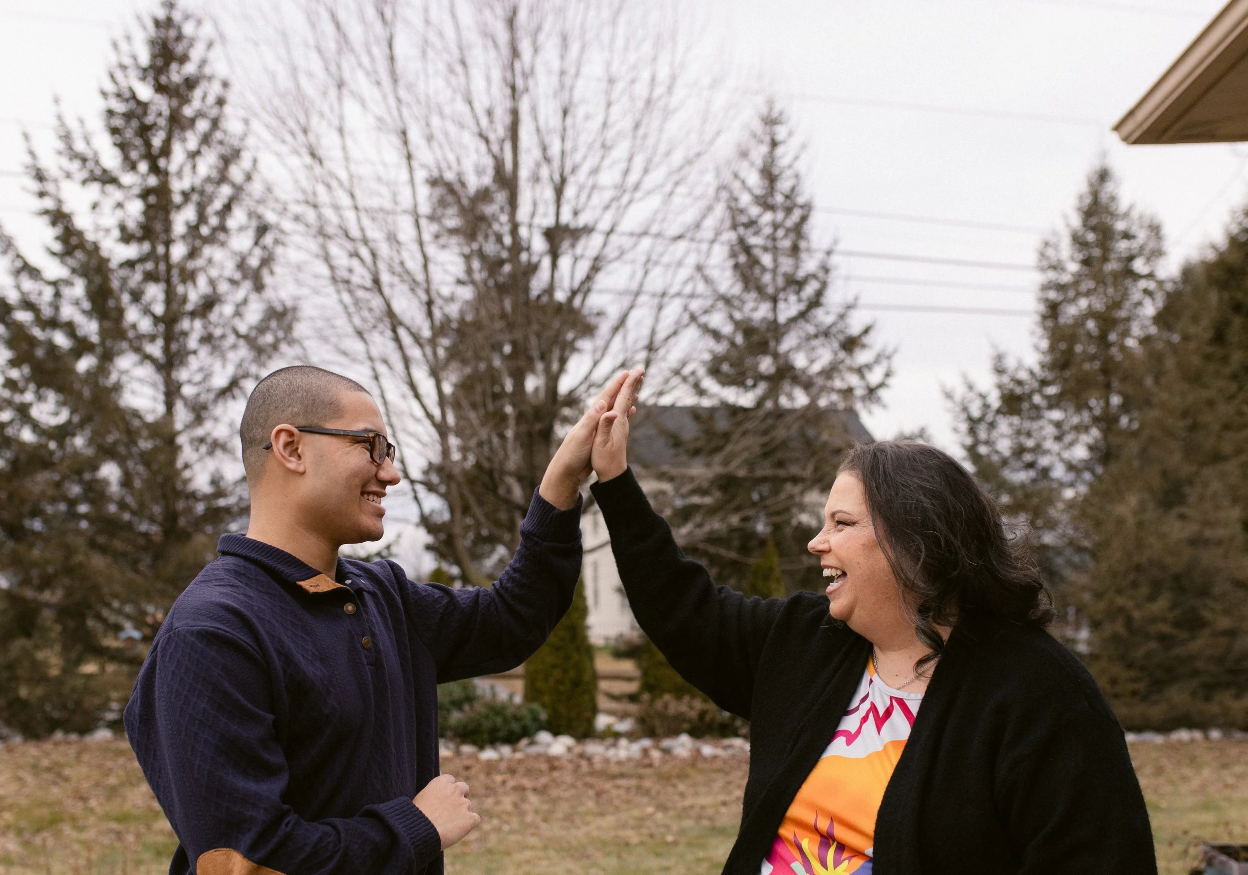 A young man and a woman exchange high-fives outdoors in a yard, both smiling and facing each other with trees and a house in the background during overcast weather.