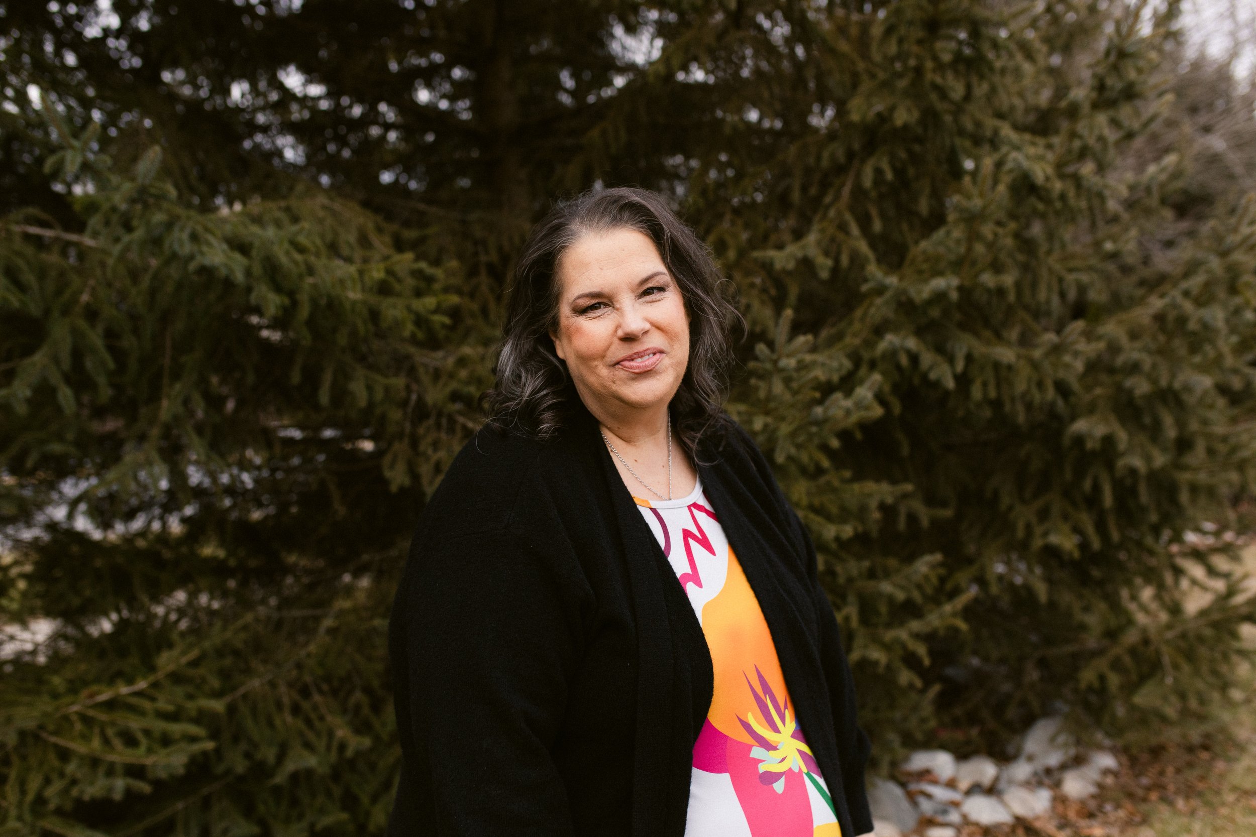 A woman with black hair wearing a black jacket and a colorful dress standing outdoors in front of large evergreen trees.