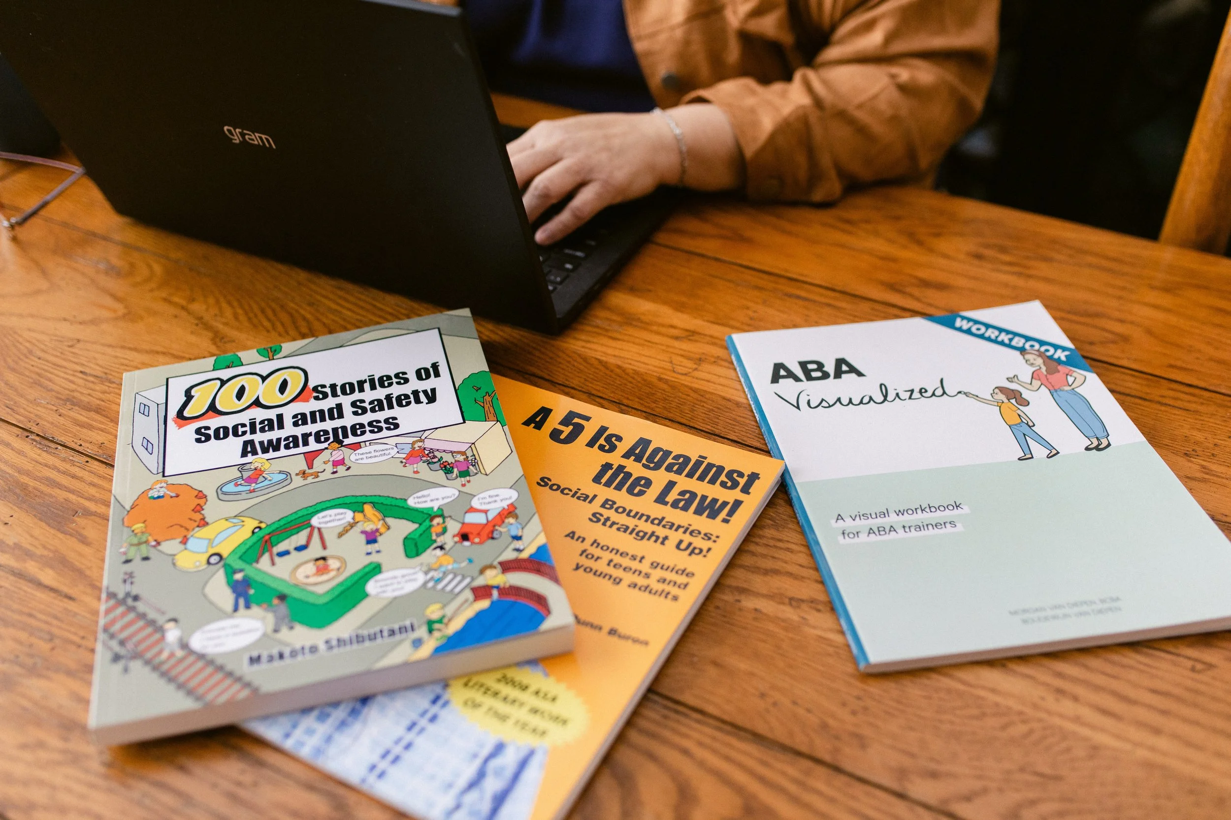 Books and a laptop on a wooden table, including a book titled '100 Stories of Social and Safety Awareness,' a workbook titled 'ABA Visualized,' and a pamphlet titled 'A 5 Is Against the Law!'