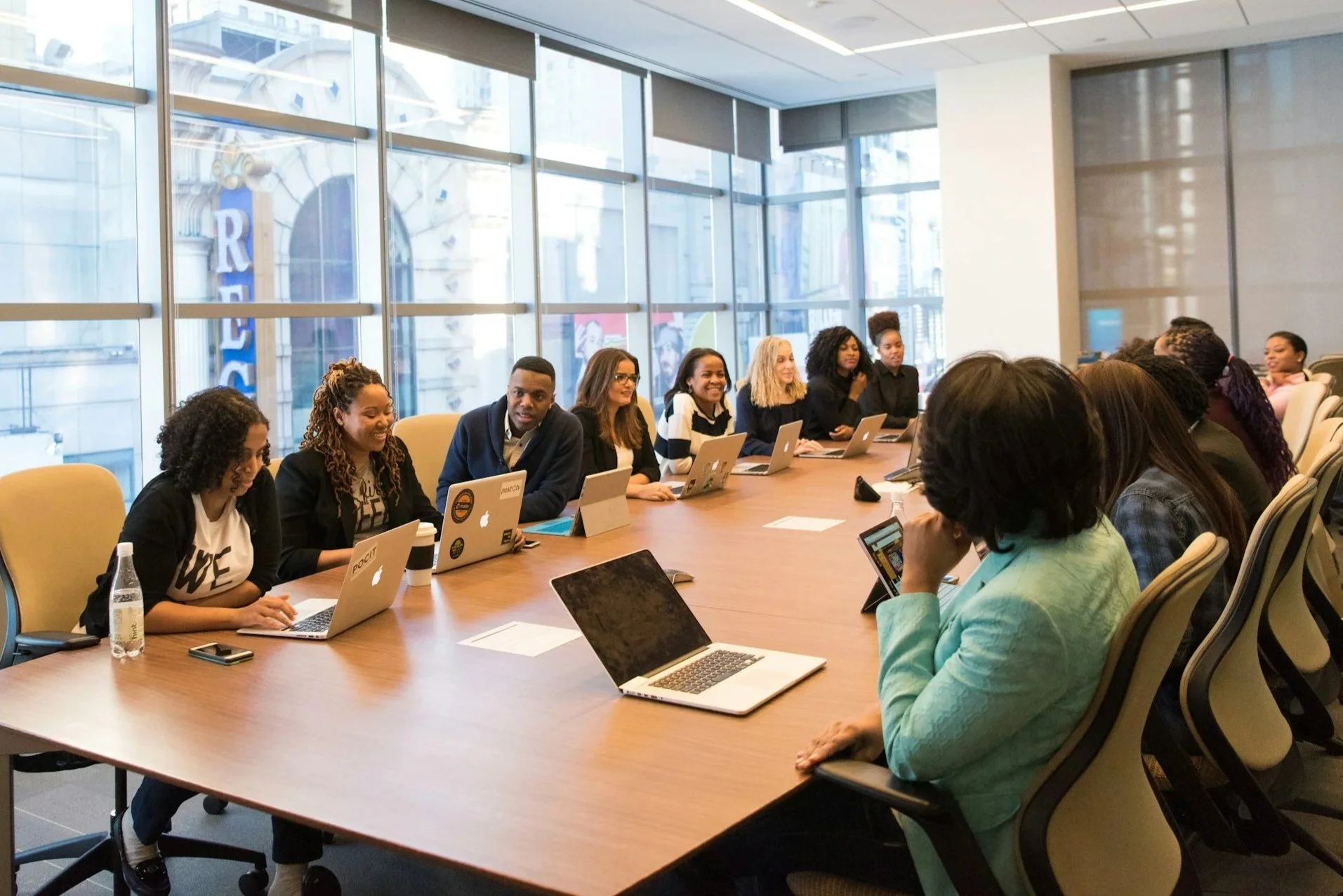 A diverse group of people sitting around a large table in a modern conference room with floor-to-ceiling windows, working on laptops and engaging in a discussion.