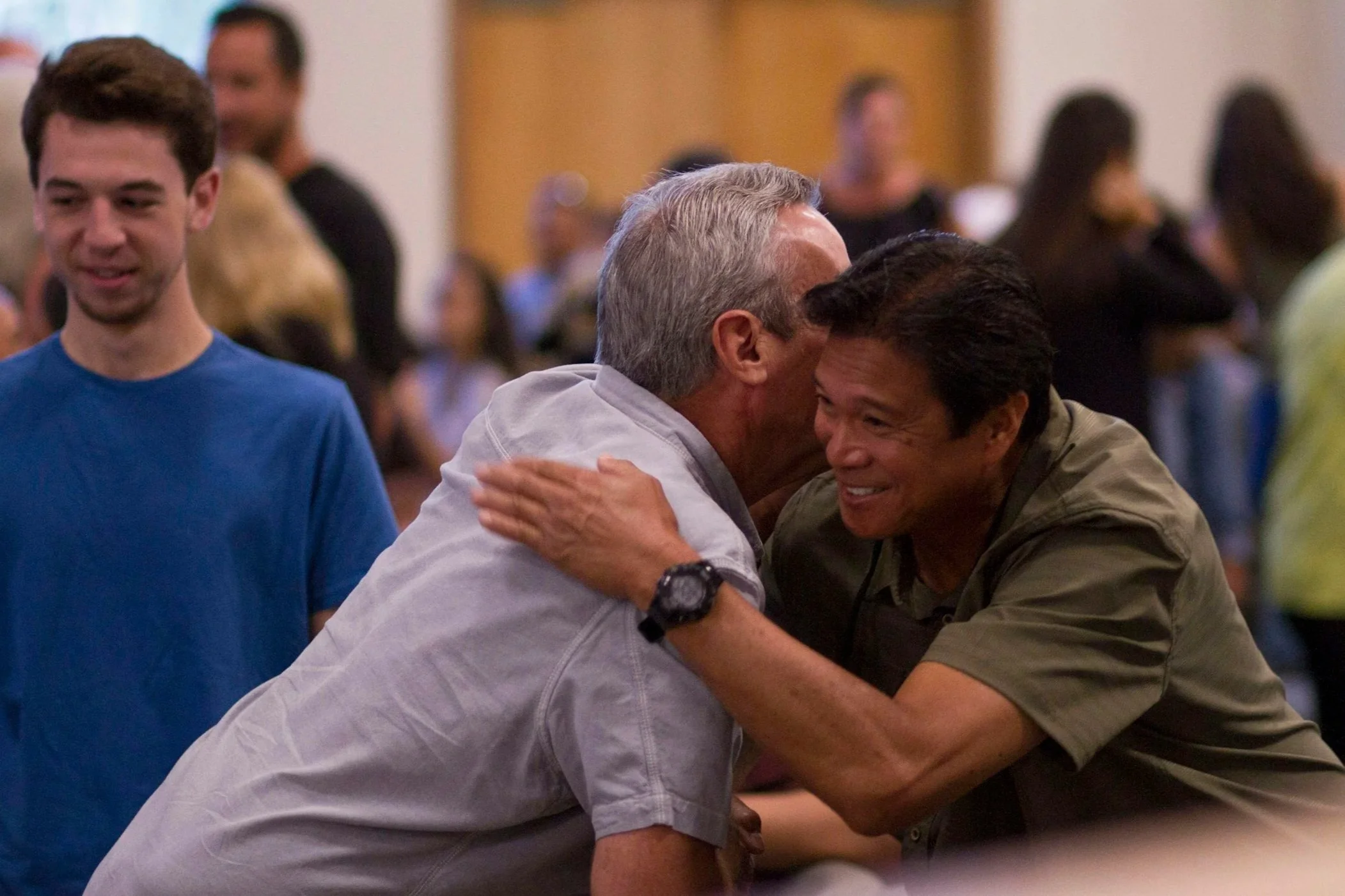Two men hugging and smiling at a gathering, with people in the background.