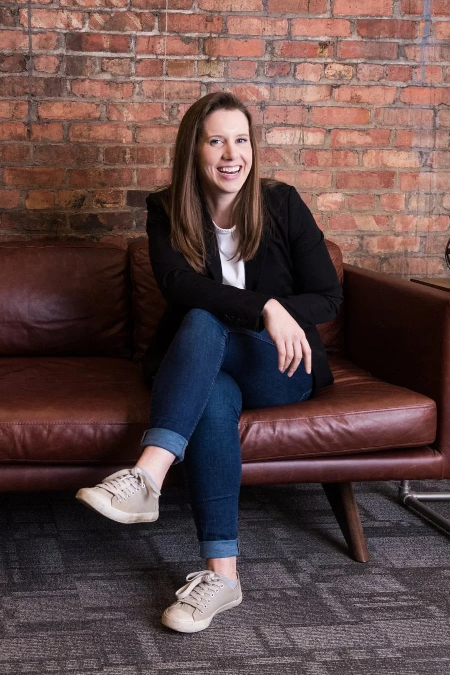 A woman with long brown hair, wearing a black blazer, white shirt, blue jeans, and beige sneakers, sitting on a brown leather sofa against a brick wall, smiling with one knee crossed over the other.