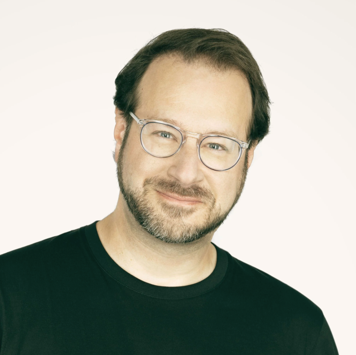 A man with glasses, a beard, and short brown hair, wearing a black shirt, smiling against a plain light background.